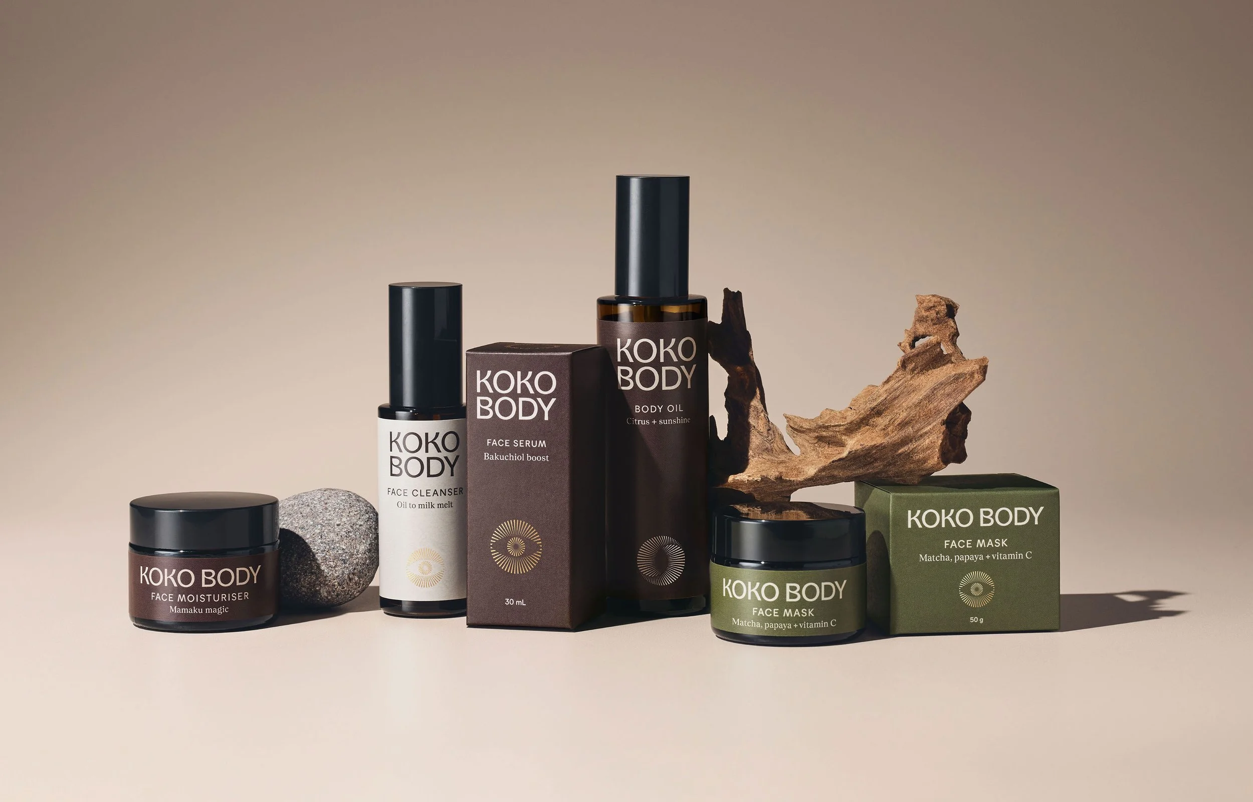 KOKO Body skincare products including face moisturizer, face cleanser, face serum, body oil, and face masks arranged on a neutral background with a piece of driftwood and stone.