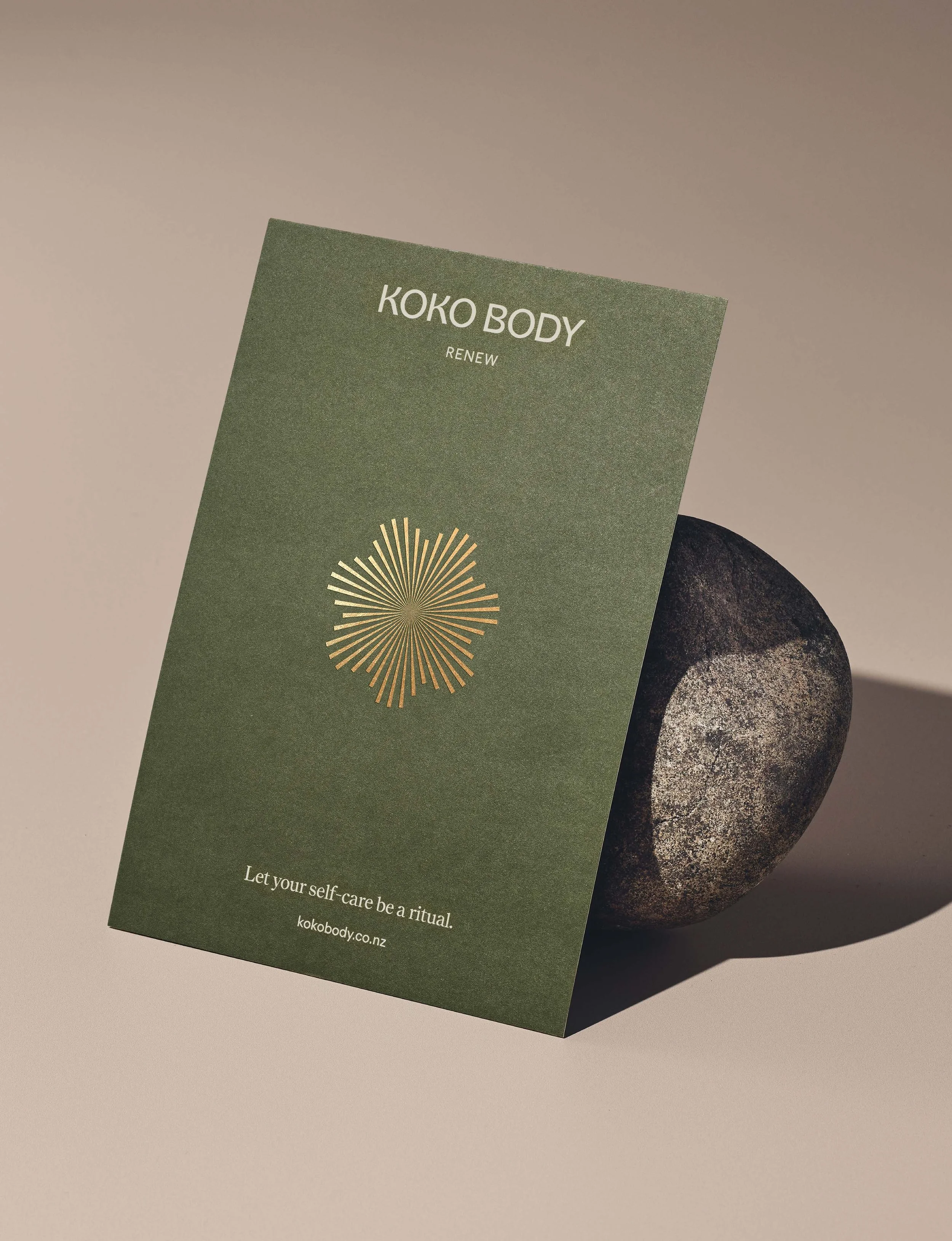 Green promotional flyer for Koko Body with sleek gold logo and text encouraging self-care ritual, resting on a smooth stone with shadow.