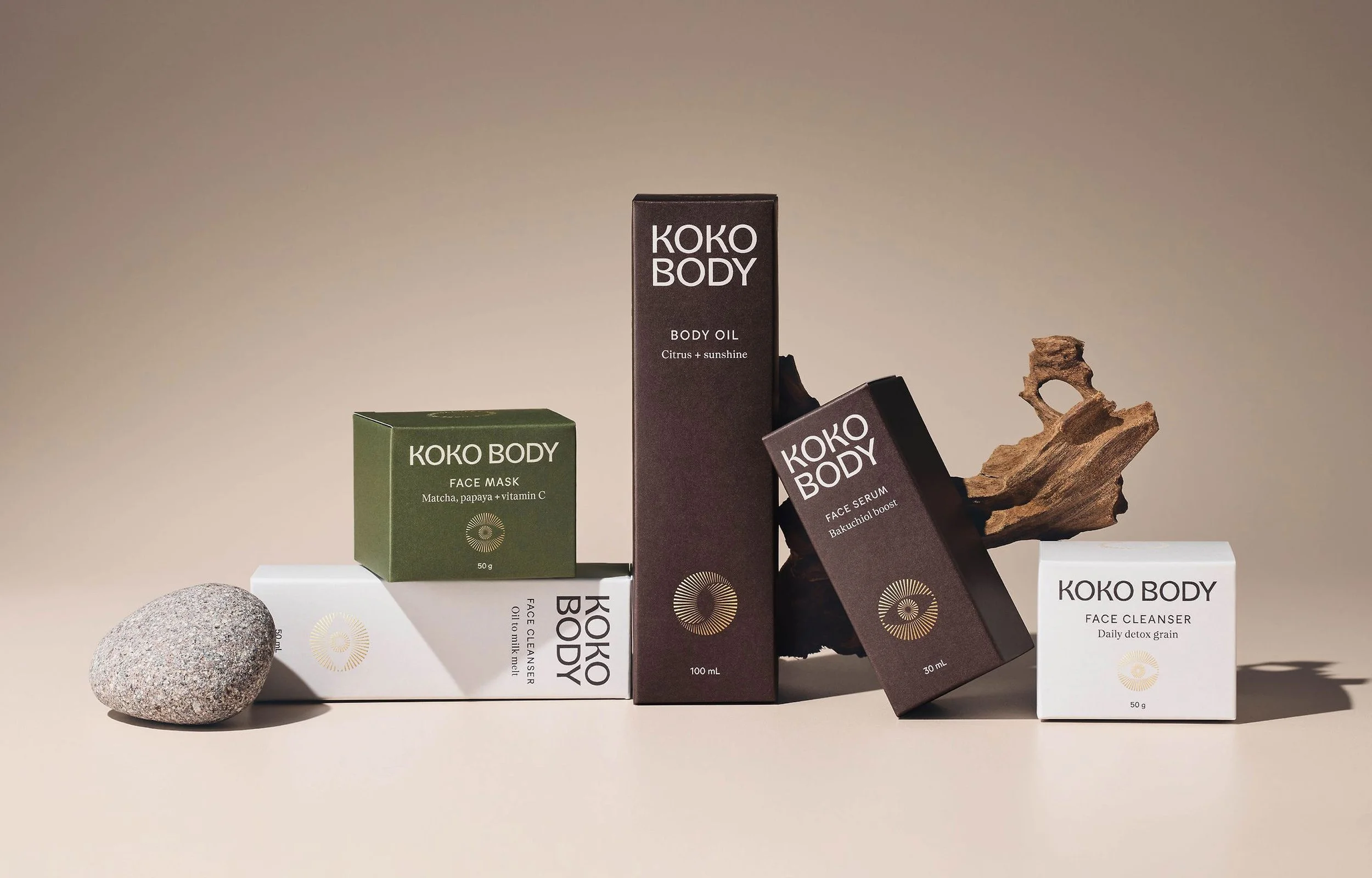 Collection of KOKO BODY skincare products including face mask, face cleanser, face serum and body oil arranged on a beige surface with a rock and a piece of driftwood.