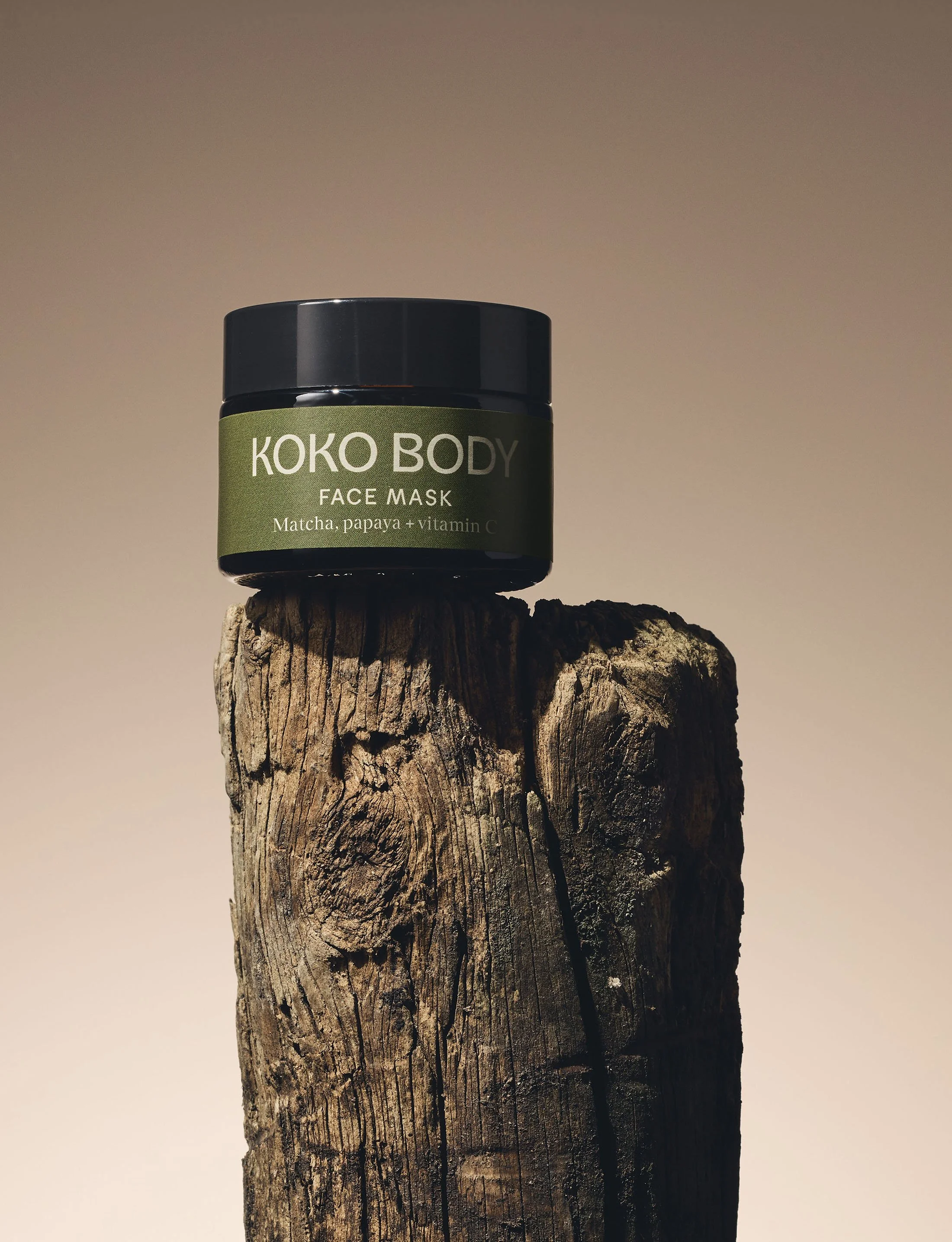 A black jar of KOKO BODY face mask with green label, placed on top of a weathered wooden post against a plain background.