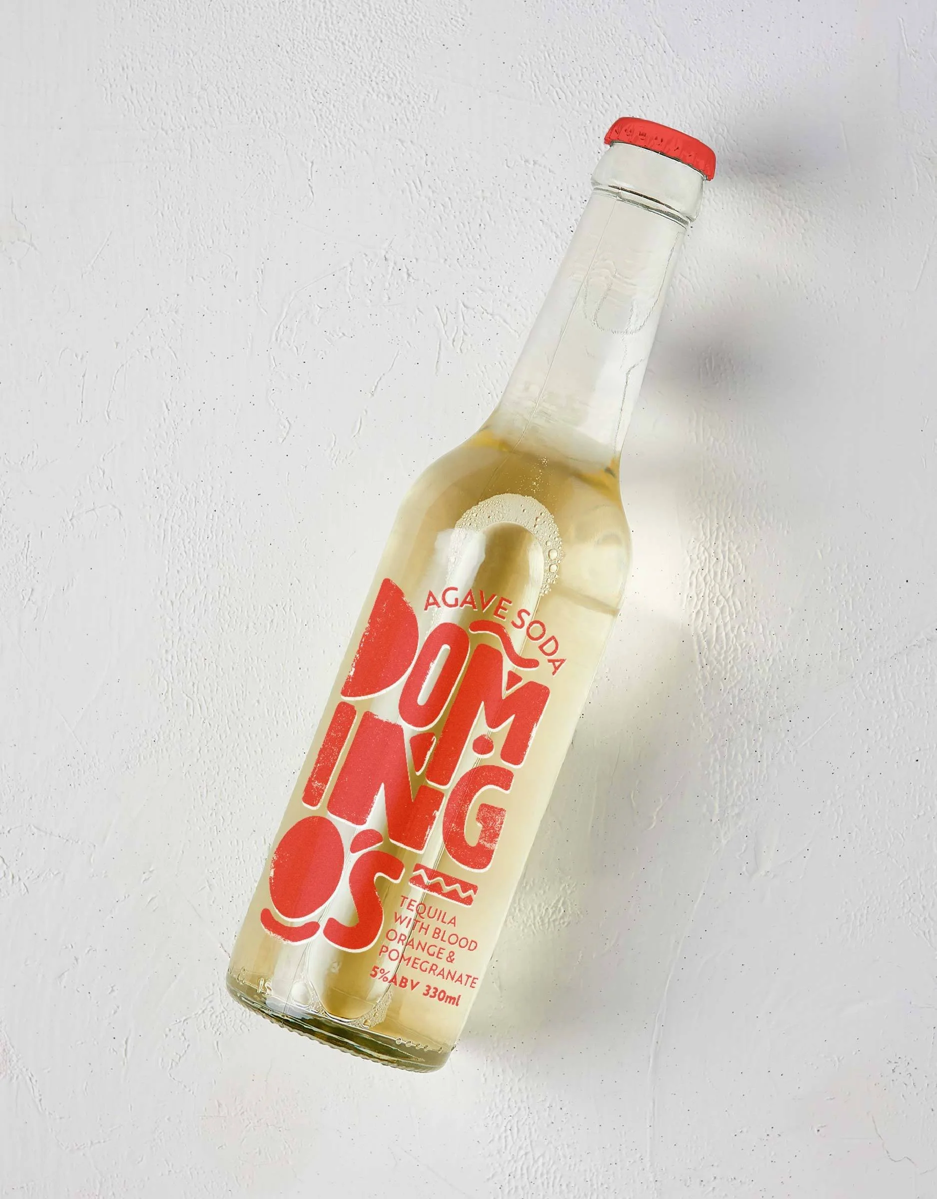 A clear glass bottle of agave soda with a red cap, containing a yellow beverage, tilted upward against a white textured background, featuring a red label that says "Domingos" and notes the drink contains tequila with blood orange and pomegranate.