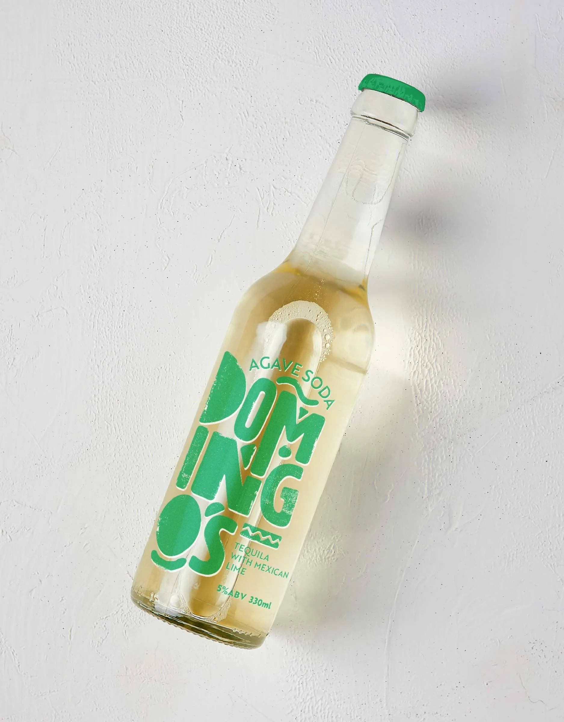A glass bottle of agave soda with a green cap, labeled with green and white text, containing a light-colored fizzy beverage, on a white textured surface.