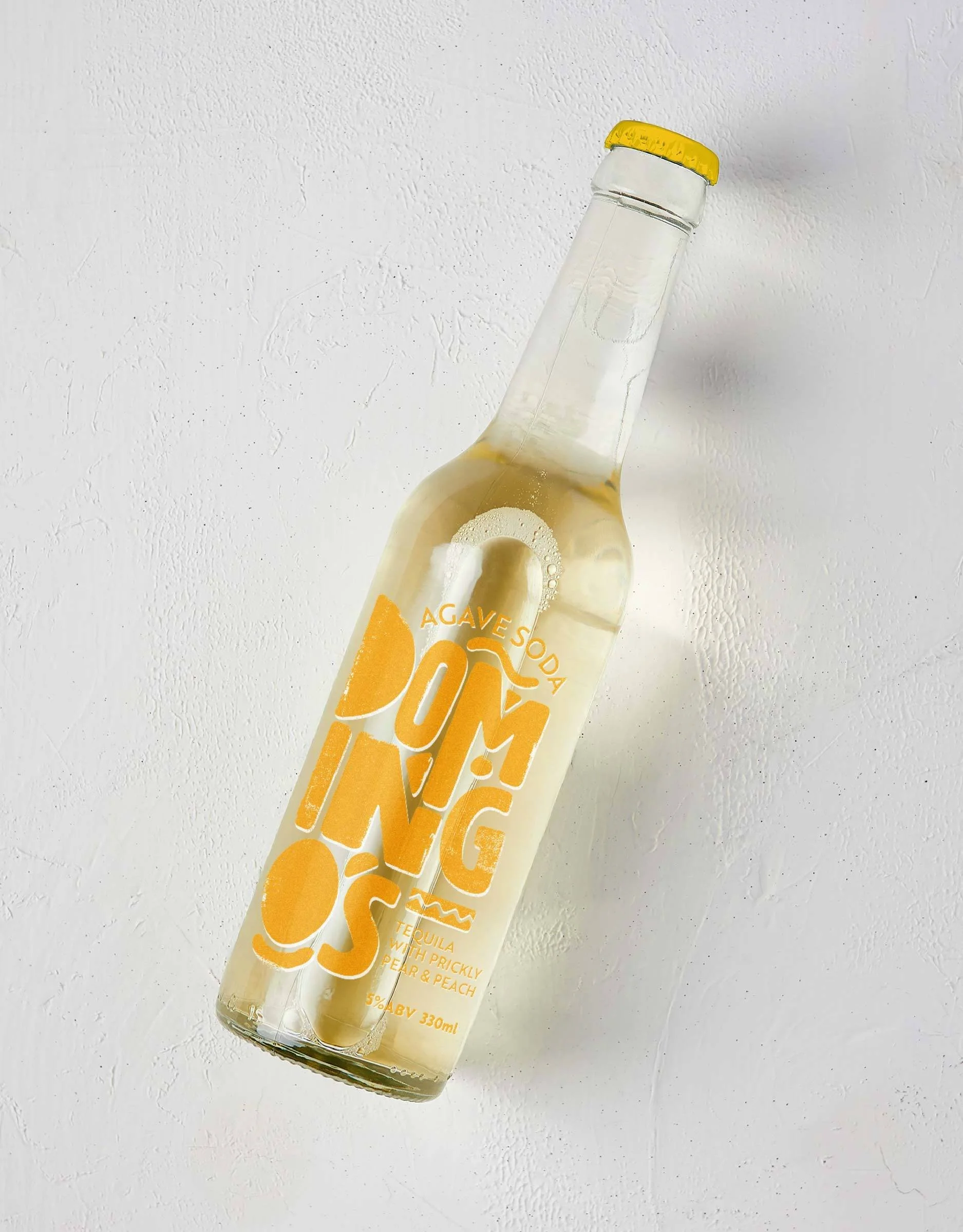 A clear glass bottle of agave soda with a yellow cap, labeled 'Pomping' with flavor notes of prickly pear and peach, on a white textured surface.