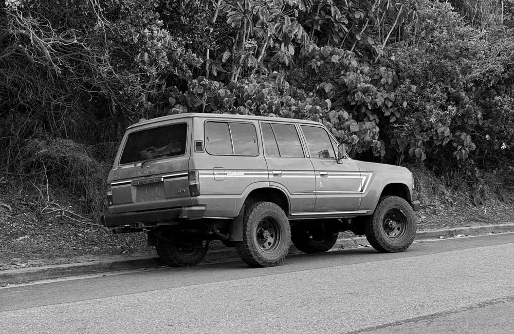 Black and white photo of a toyota landcruiser FJ60 parked on the side of the road with bushes behind it.