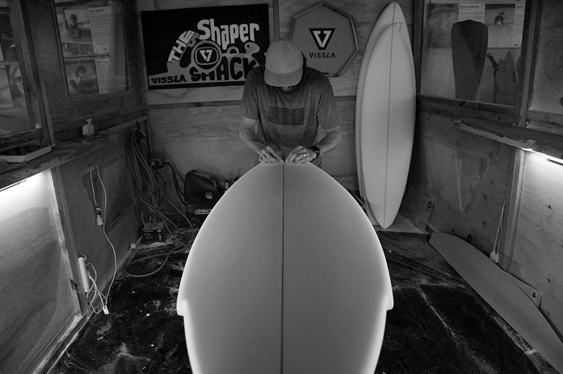 A person working on shaping a surfboard in a workshop with surfboards and banners in the background.