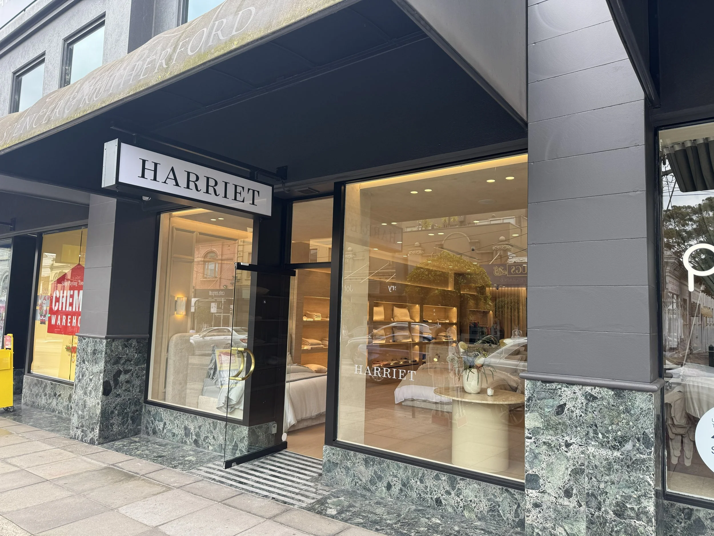 Storefront with the sign 'HARRIET', large glass windows showing beds and home decor inside, dark gray exterior with marble accents, sidewalk in front.
