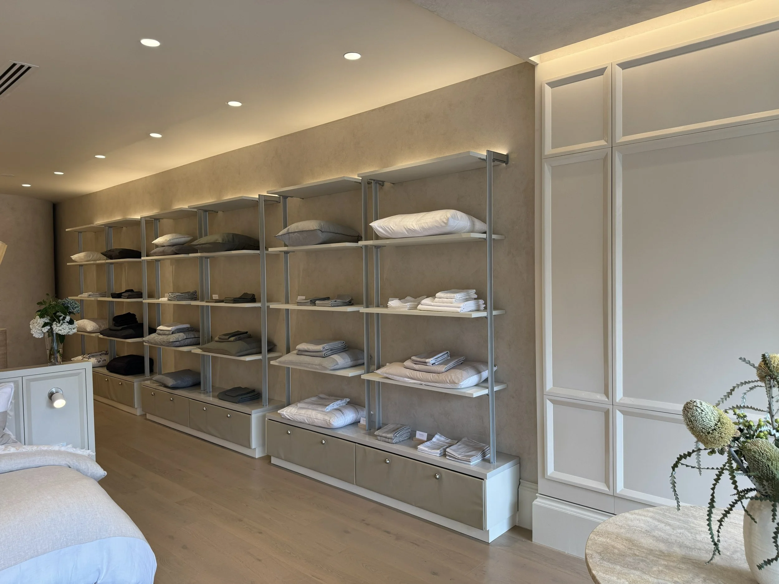 Modern bedroom with shelves displaying neatly folded towels, pillows, and bedding, beige walls, white panel accents, and a flower arrangement on a table.