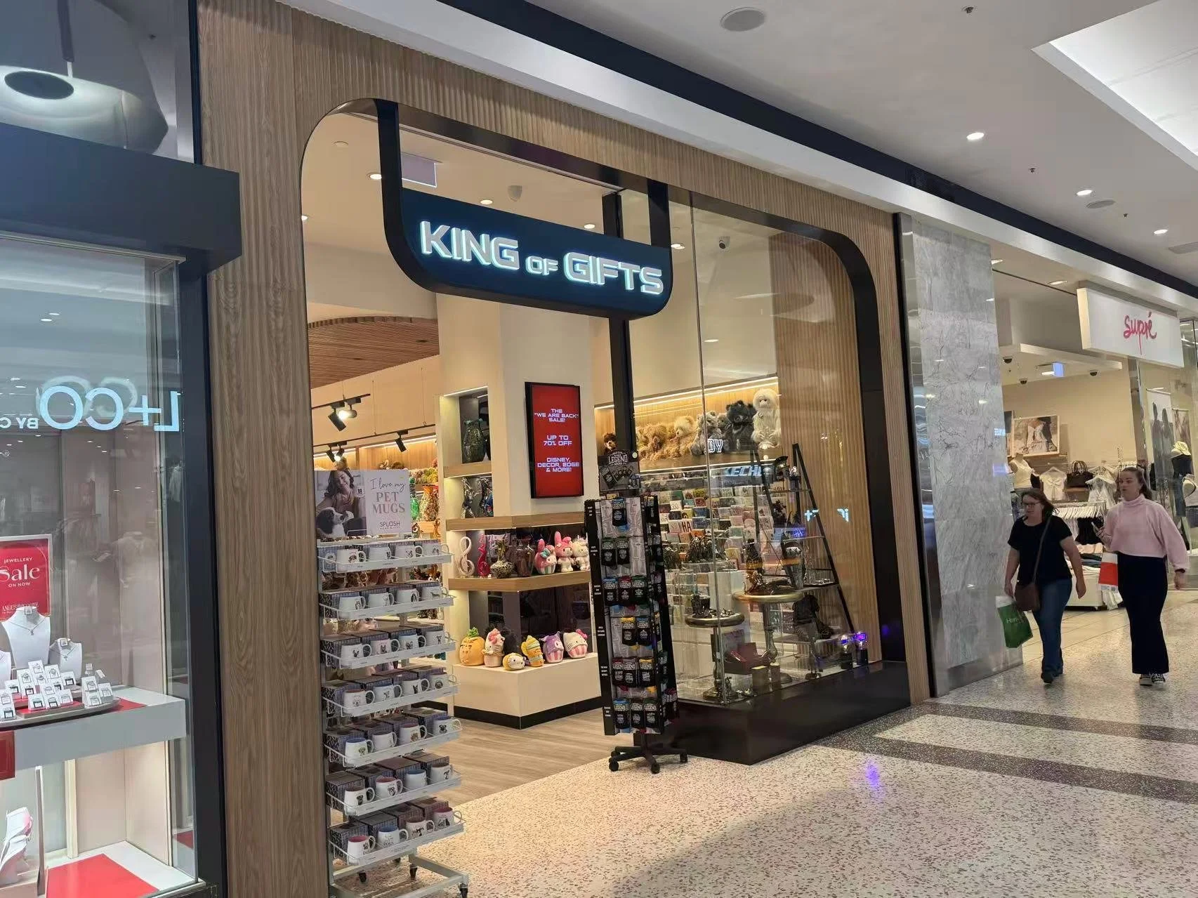Storefront of 'King of Gifts' in a shopping mall, with displays of plush toys, mugs, and accessories for sale, and two women walking past outside.