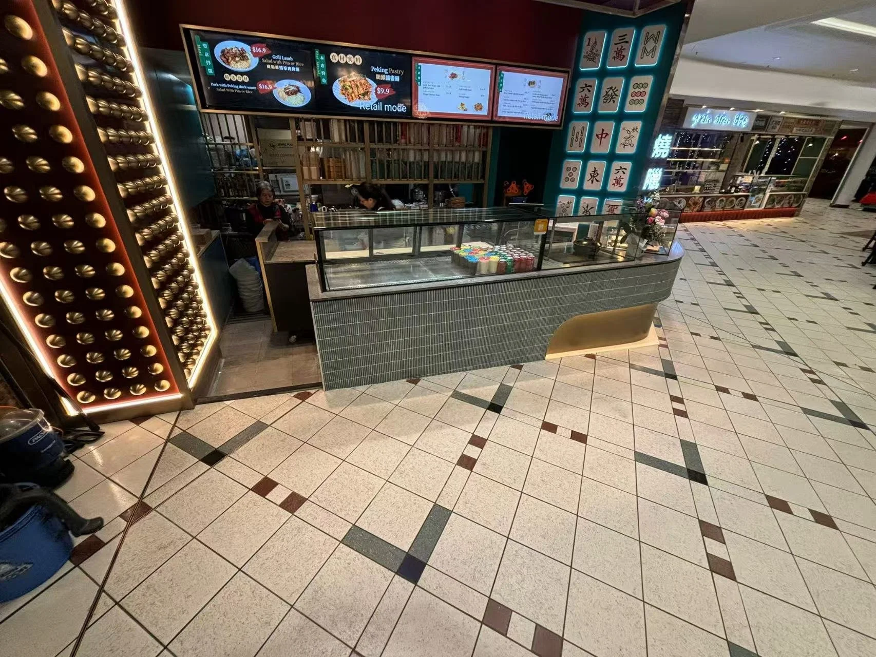 A small food counter at a shopping mall, with digital menu screens displaying Asian dishes and prices, behind a glass display case, with two employees working behind the counter.