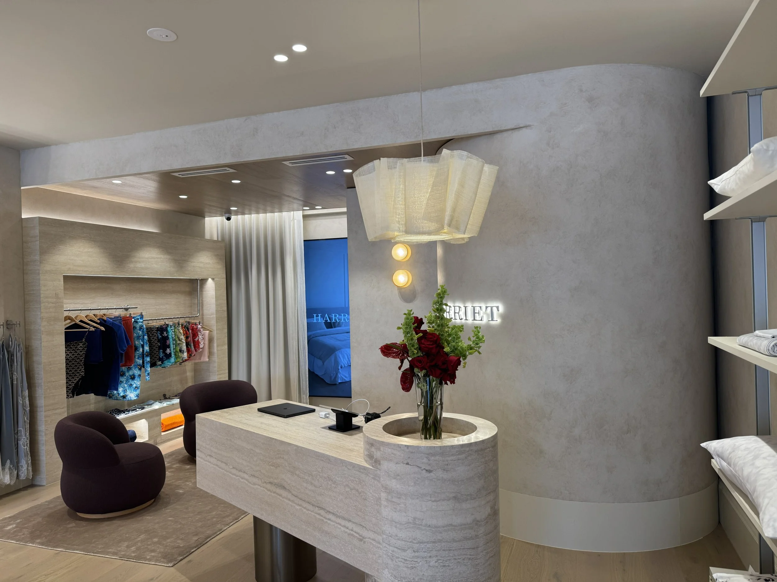 Interior of a retail boutique with clothing display, a curved wall with backlit sign, a table with a flower arrangement, and a bedroom visible through a doorway.