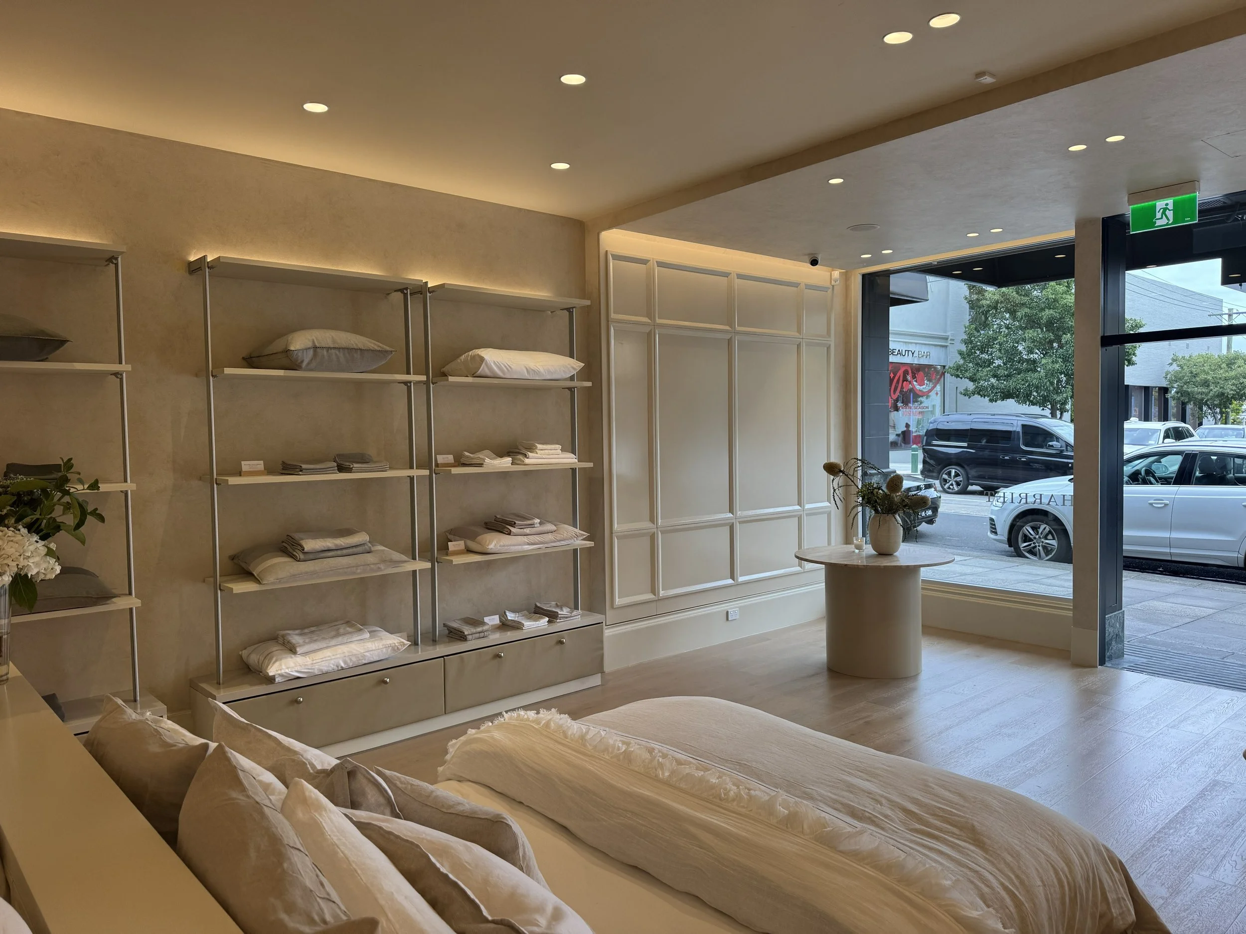 Interior of a minimalist bedroom store with shelves of neatly folded linens and pillows, a round table with a flower vase, and a large window showing parked cars outside.