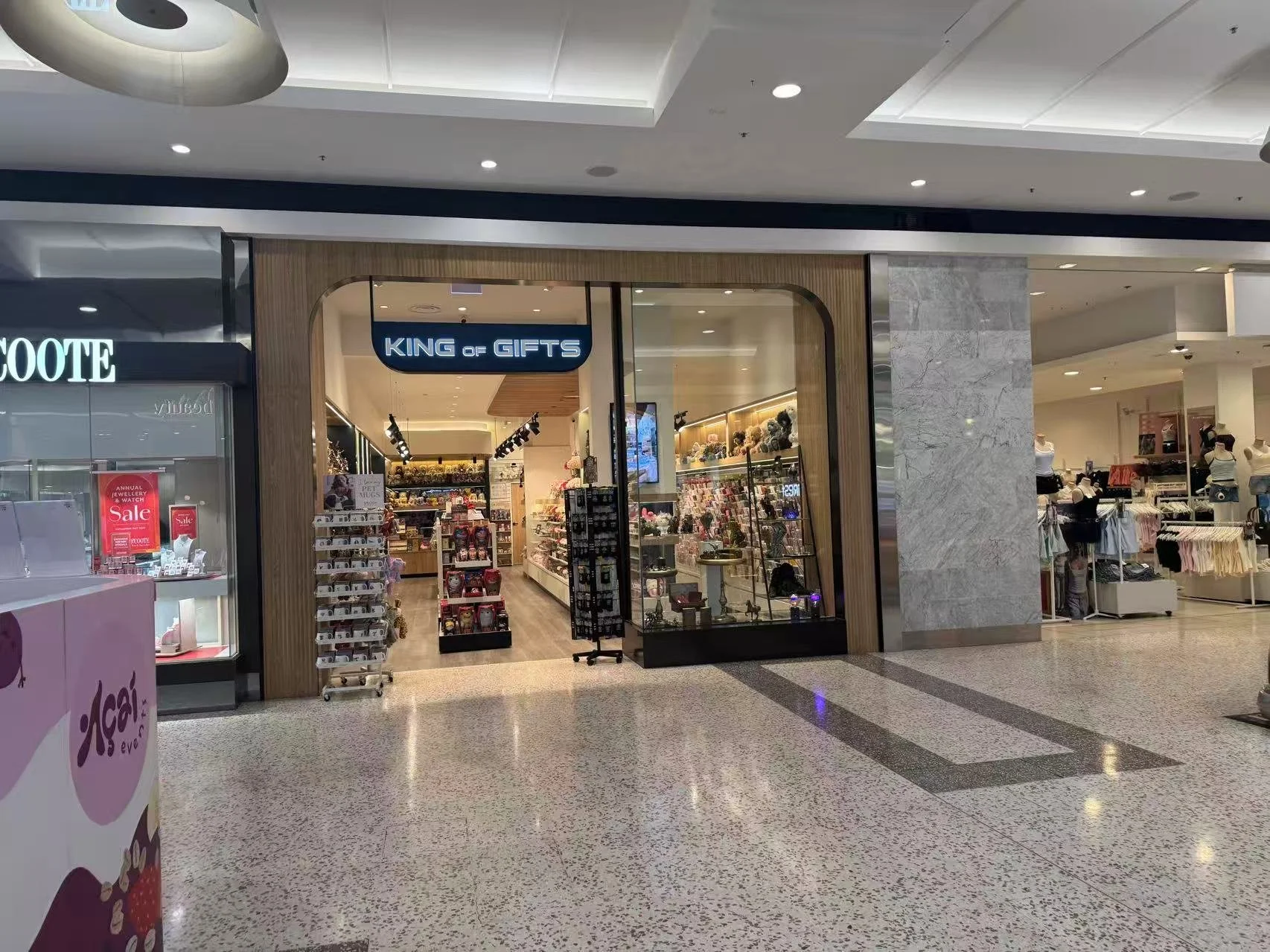 Entrance to a gift shop called 'King of Gifts' inside a mall. The shop has shelves with various gifts and accessories, and a digital sign displaying prices.
