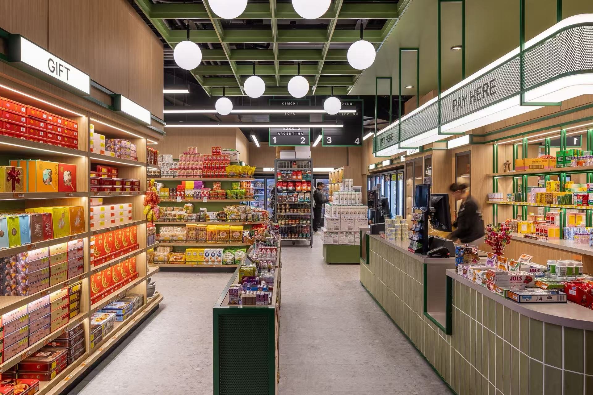 Interior of a modern convenience store with shelves stocked with colorful packaged goods, checkout counter on the right, and pay here signs overhead.