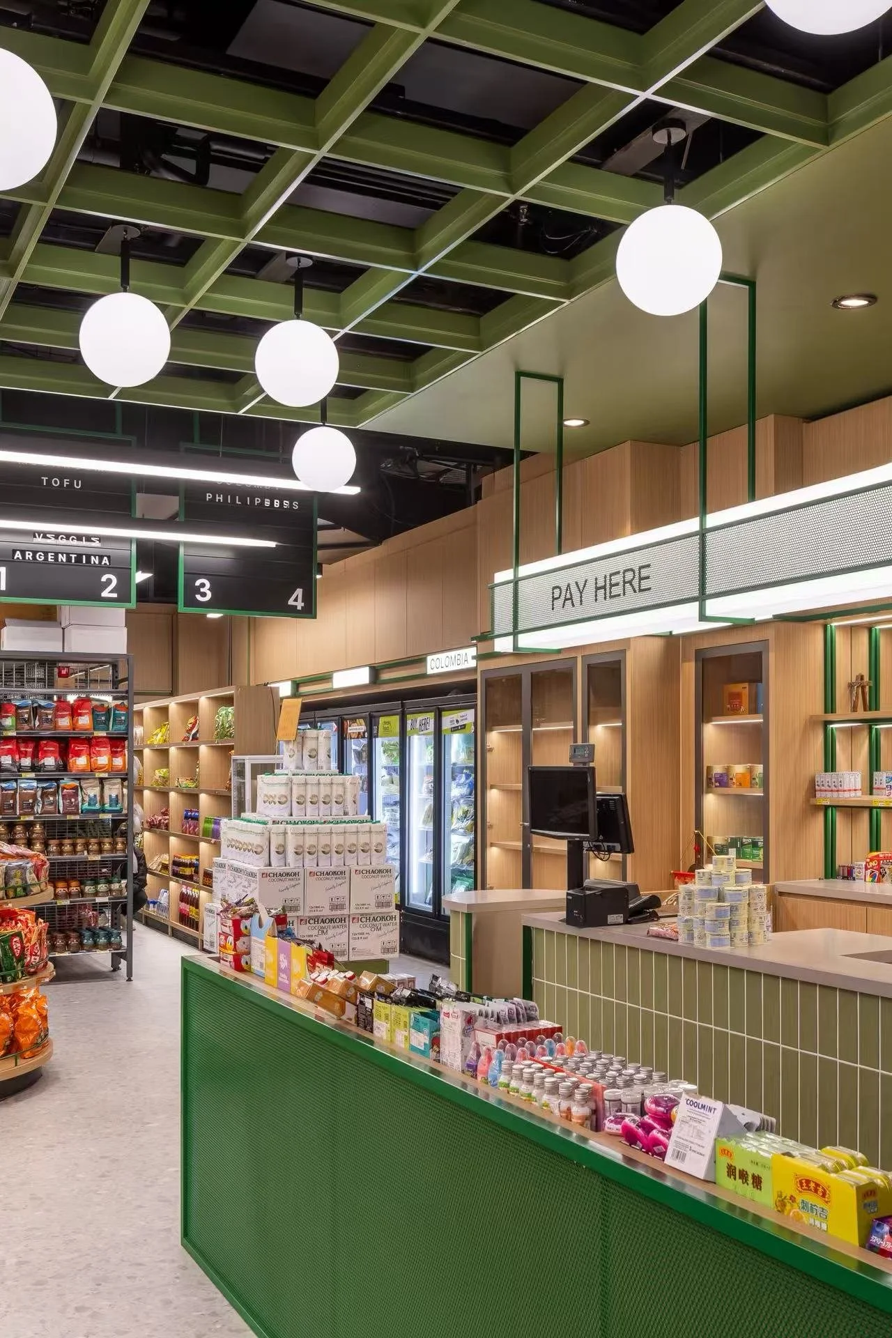 Inside a modern grocery store or convenience shop with a checkout counter that says "Pay Here," shelves stocked with various products, a digital display showing checkout lane information, and bright, round ceiling lights.
