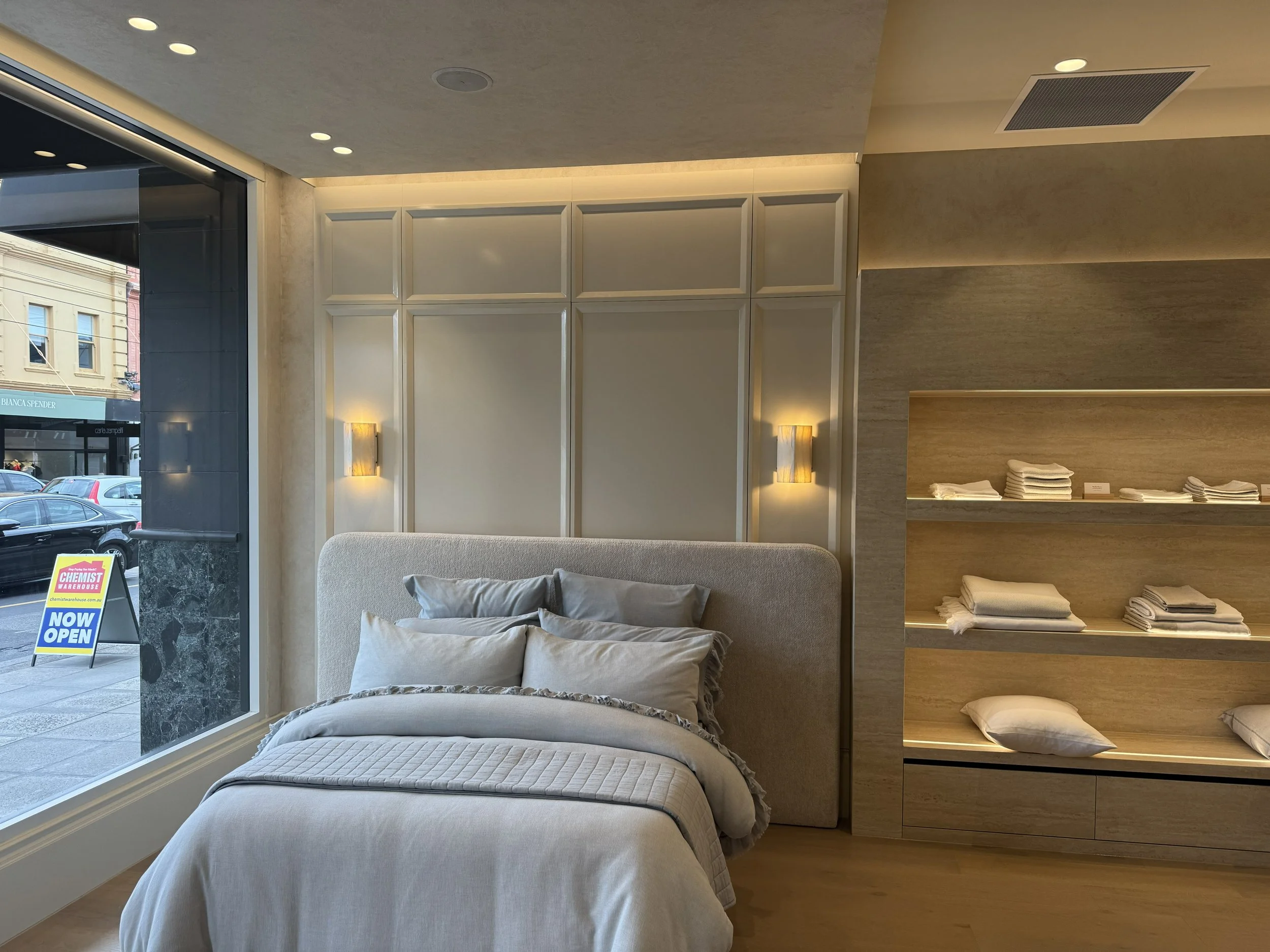 A modern bedroom with a large window, a bed with gray and beige pillows, a beige headboard, wall-mounted lights, and wooden shelves with neatly folded linens.
