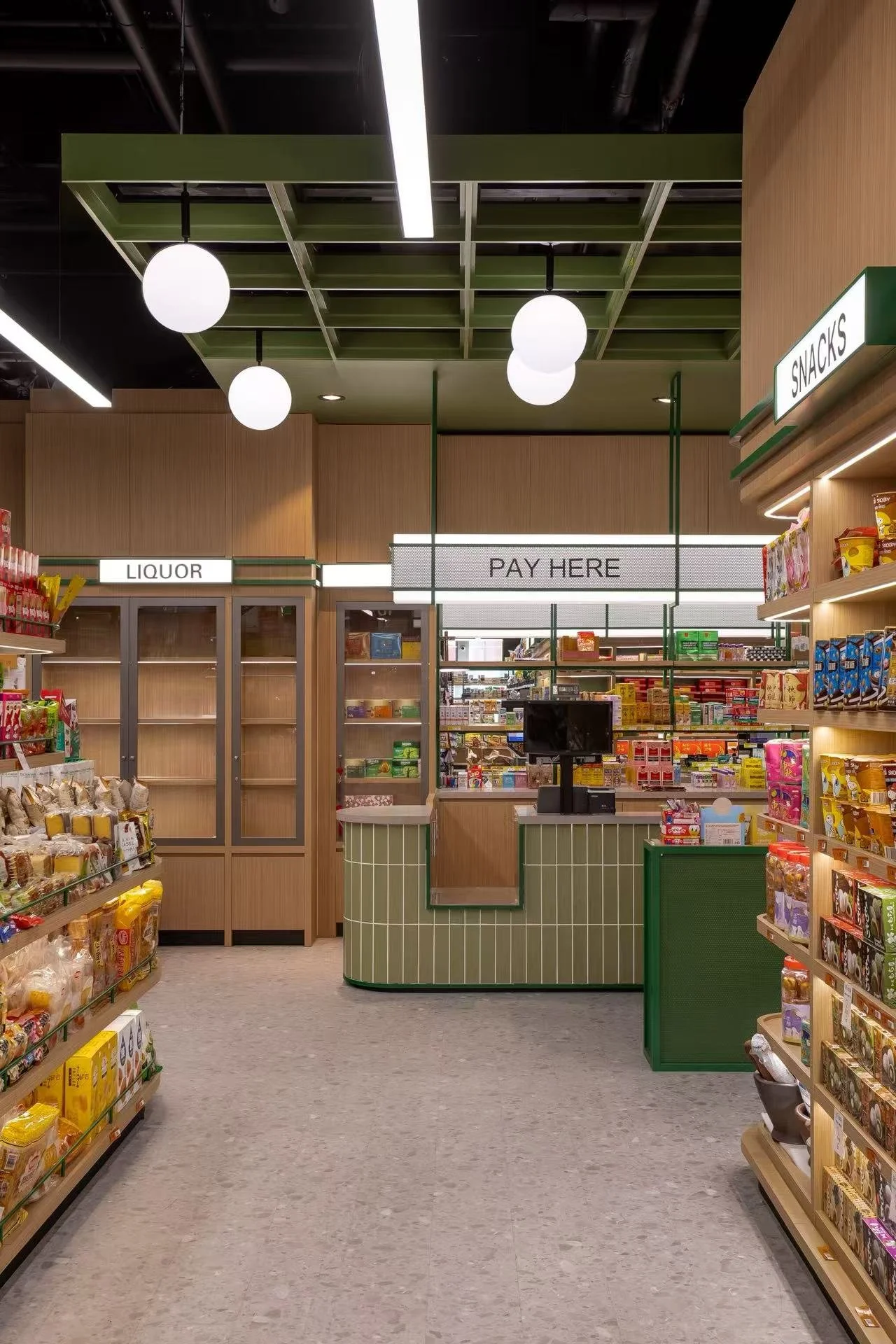 Grocery store checkout counter with signs reading 'Pay Here,' 'Liquor,' and 'Snacks,' with shelves of snack items on the right and left sides, and a green checkout kiosk in the center.