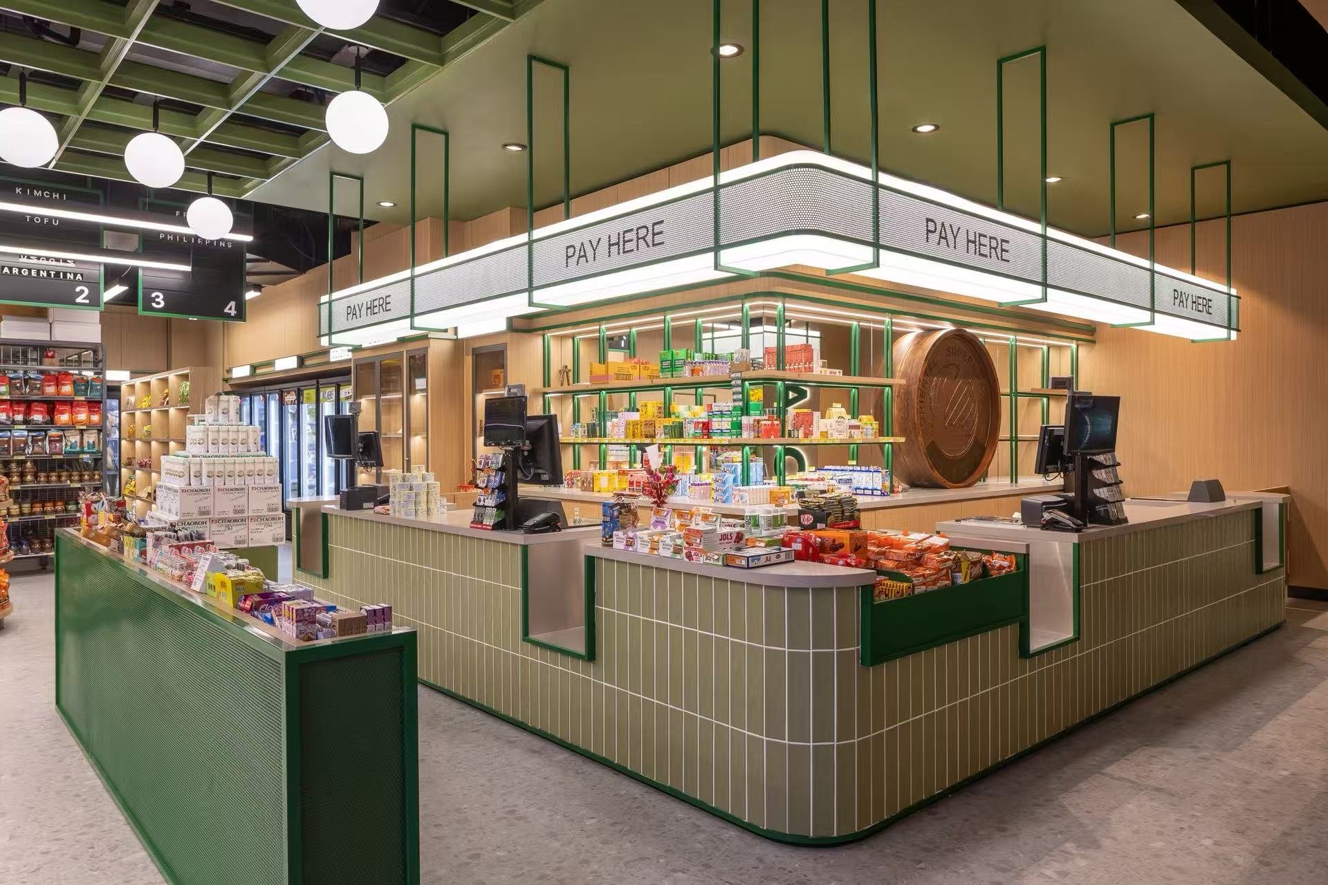 A modern checkout counter at a grocery store with a green and beige tiled front, multiple cash registers, and a large illuminated sign overhead reading 'Pay Here'. Shelves with snacks and products are visible behind the counter, along with a digital 