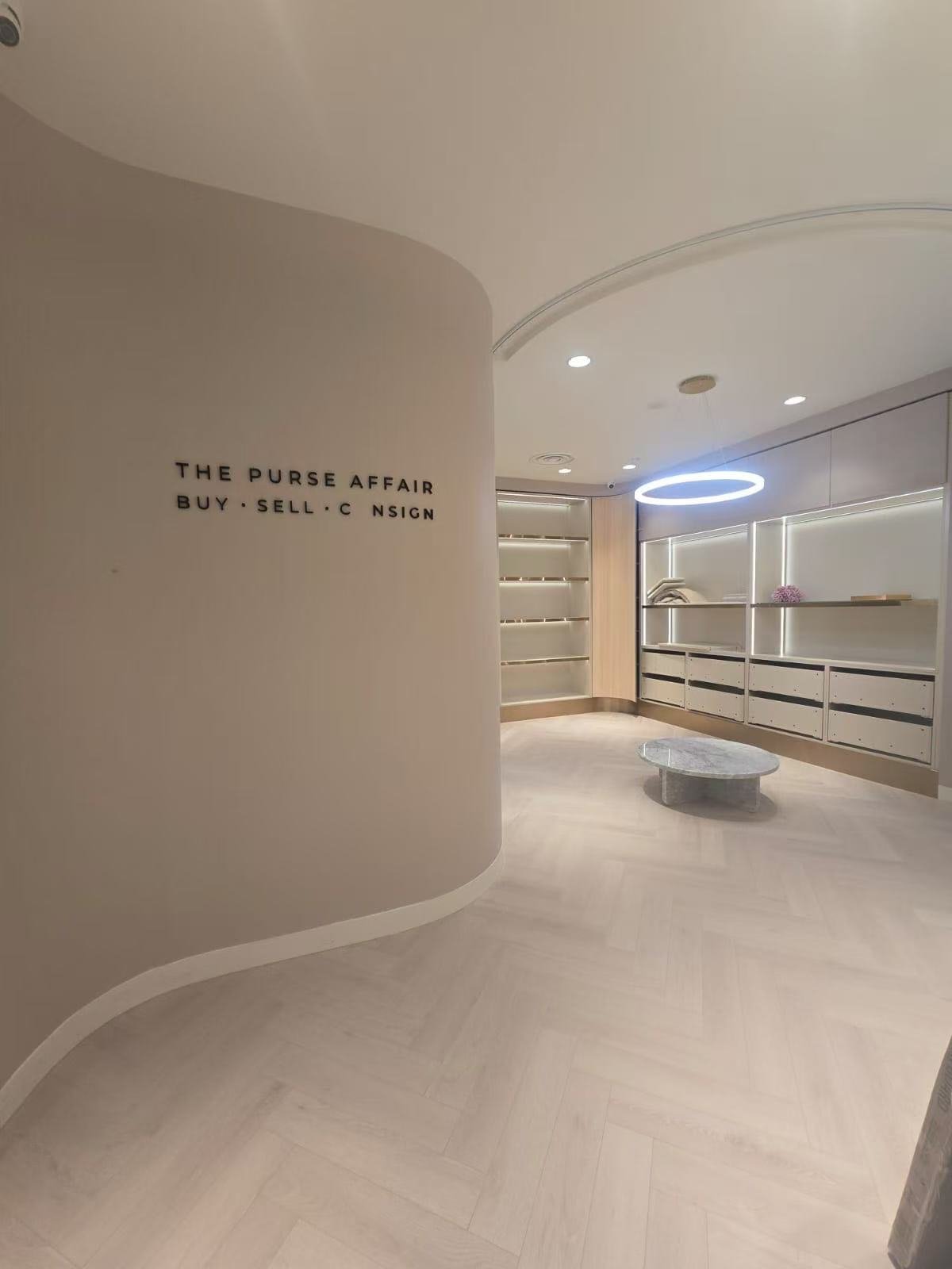Modern retail store interior with a curved wall labeled 'The Purse Affair Buy Sell Consign', minimalist shelving, a circular light fixture, and a central marble table.