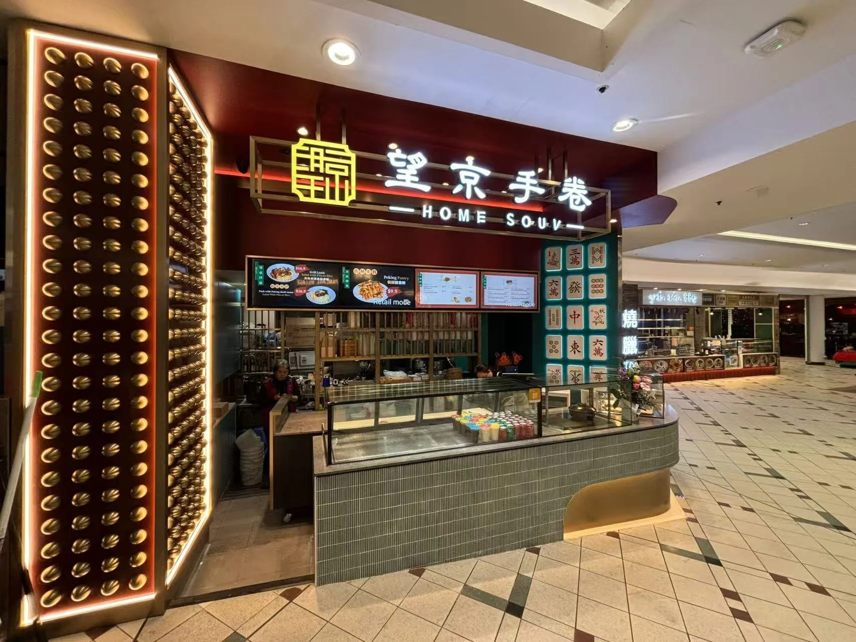 A bakery stall inside a shopping mall with a sign reading 'Home Souv' in Chinese and English. It has a glass display counter with canned goods and a cash register, surrounded by decorative elements and menu screens.