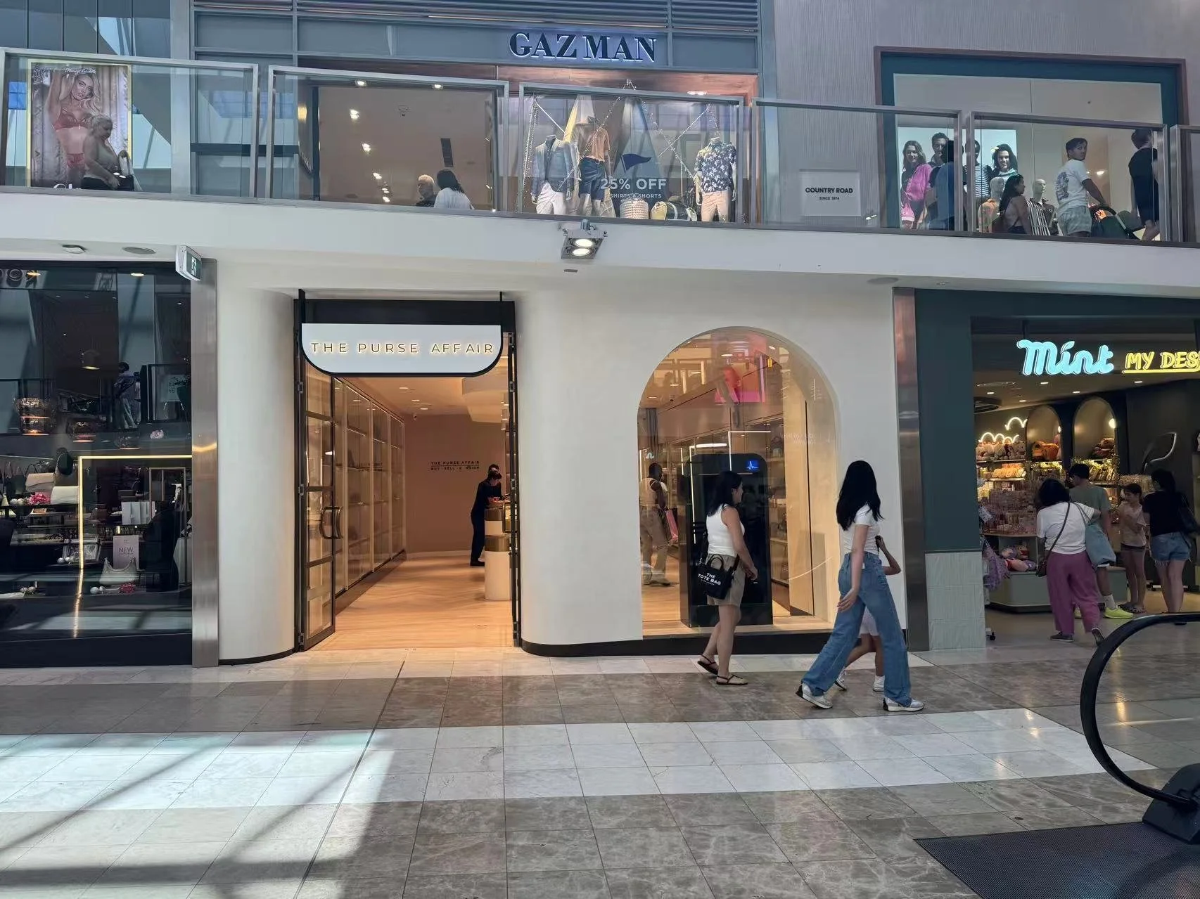 Shoppers walking outside stores in a shopping mall, including 'The Purse Affair' and 'Mint My Desk'.