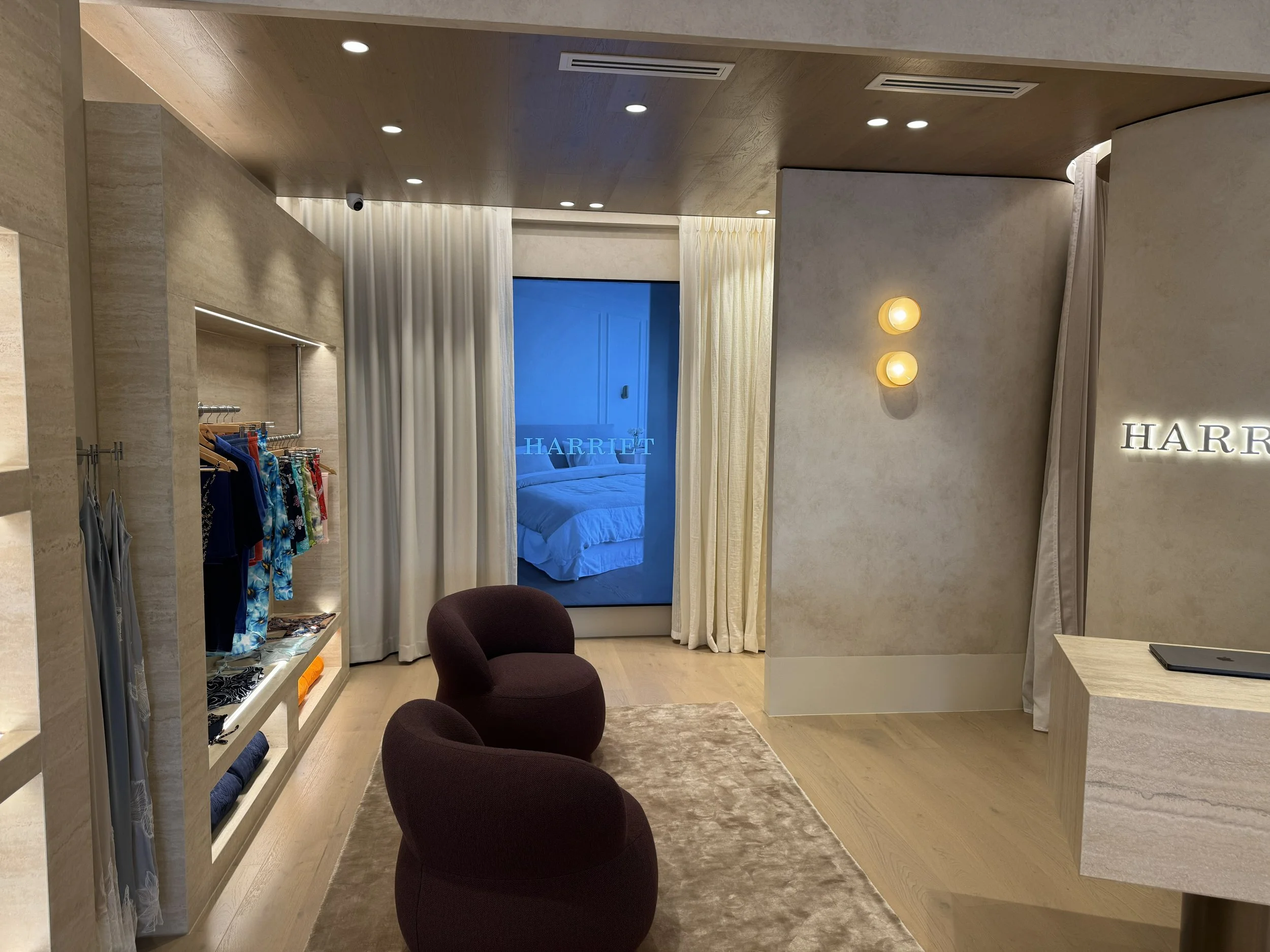 Interior of a hotel room or boutique store with clothing display, two burgundy chairs, a beige rug, and a view into a bedroom with blue-tinted lighting and a bed.