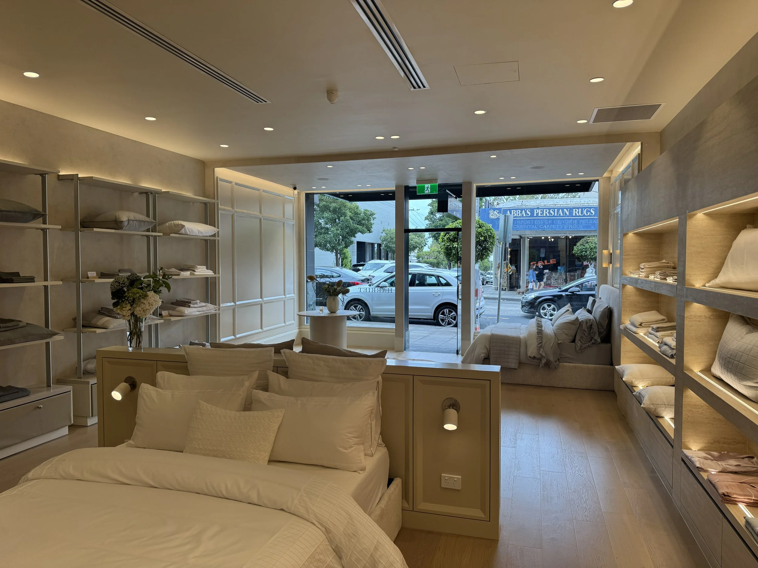 Interior of a bedding store with shelves of pillows and linens, a bed with pillows, and a seating area near large front windows looking out onto parked cars and street shops.