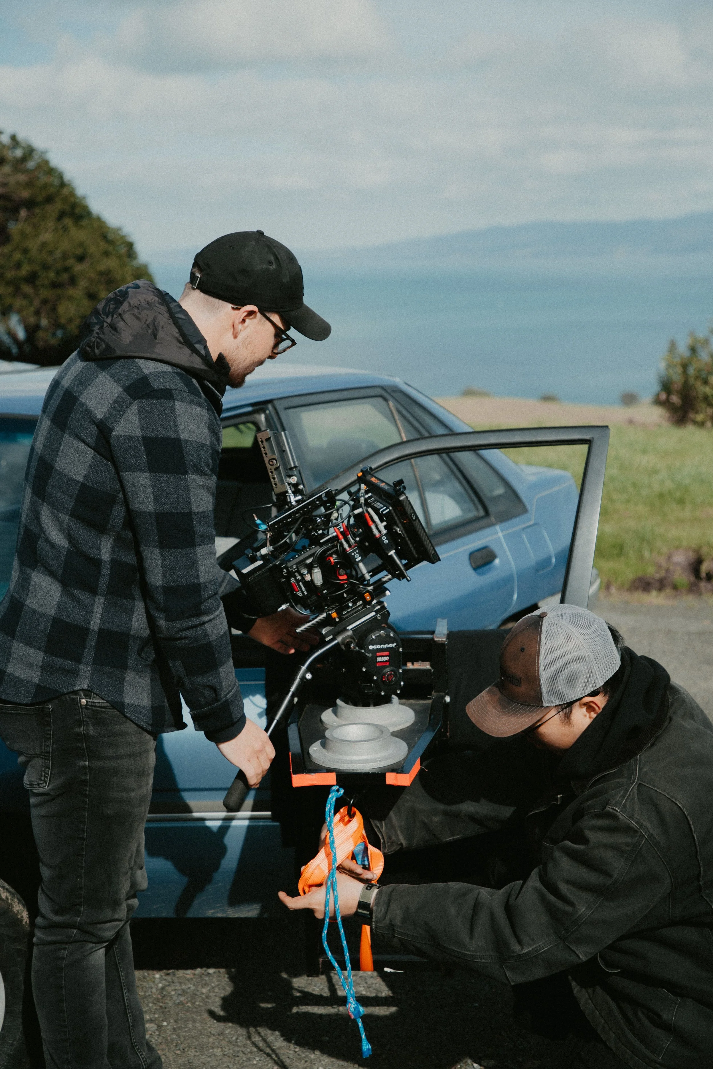 Two men working on a camera rig near a blue car outdoors, with a landscape and body of water in the background.
