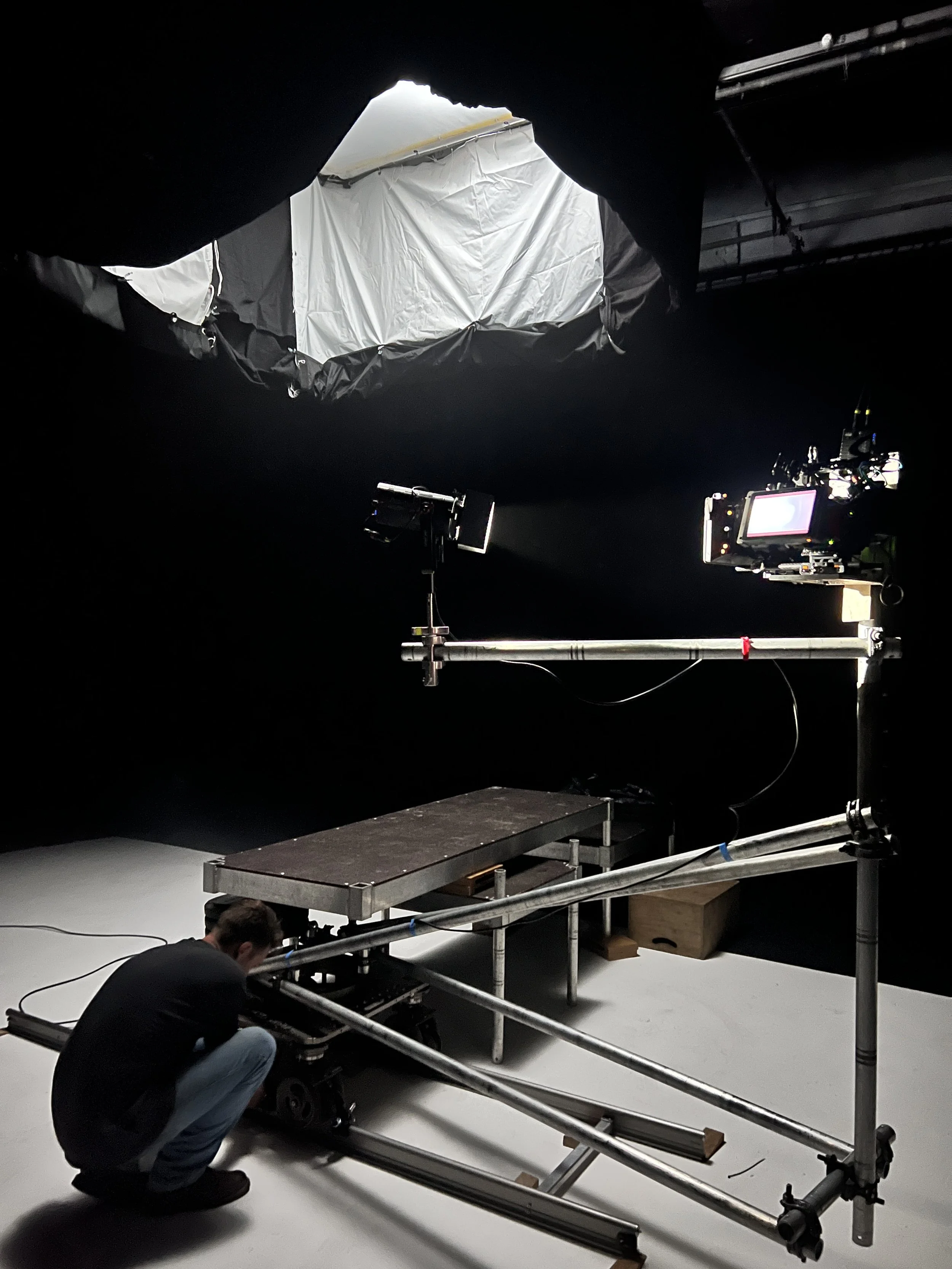 A person working on a film or photography set with equipment, a camera, and a large light source. The set is inside a black studio with a ceiling covered in black fabric and a large overhead light