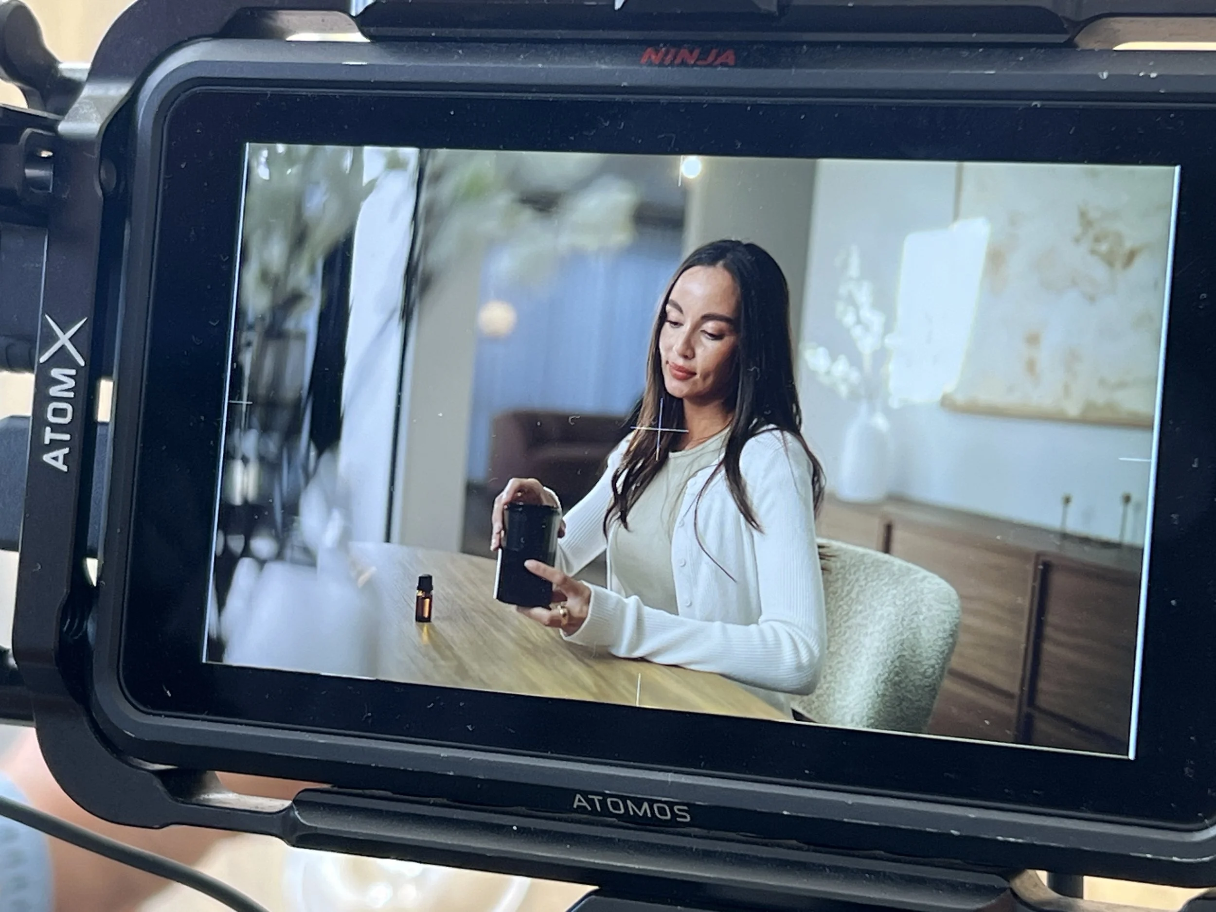 A woman with long dark hair, wearing a white cardigan, is sitting at a wooden table in a well-lit room. She is holding a small black camera and has a small bottle of essential oil or perfume on the table. The background includes a white vase and artw