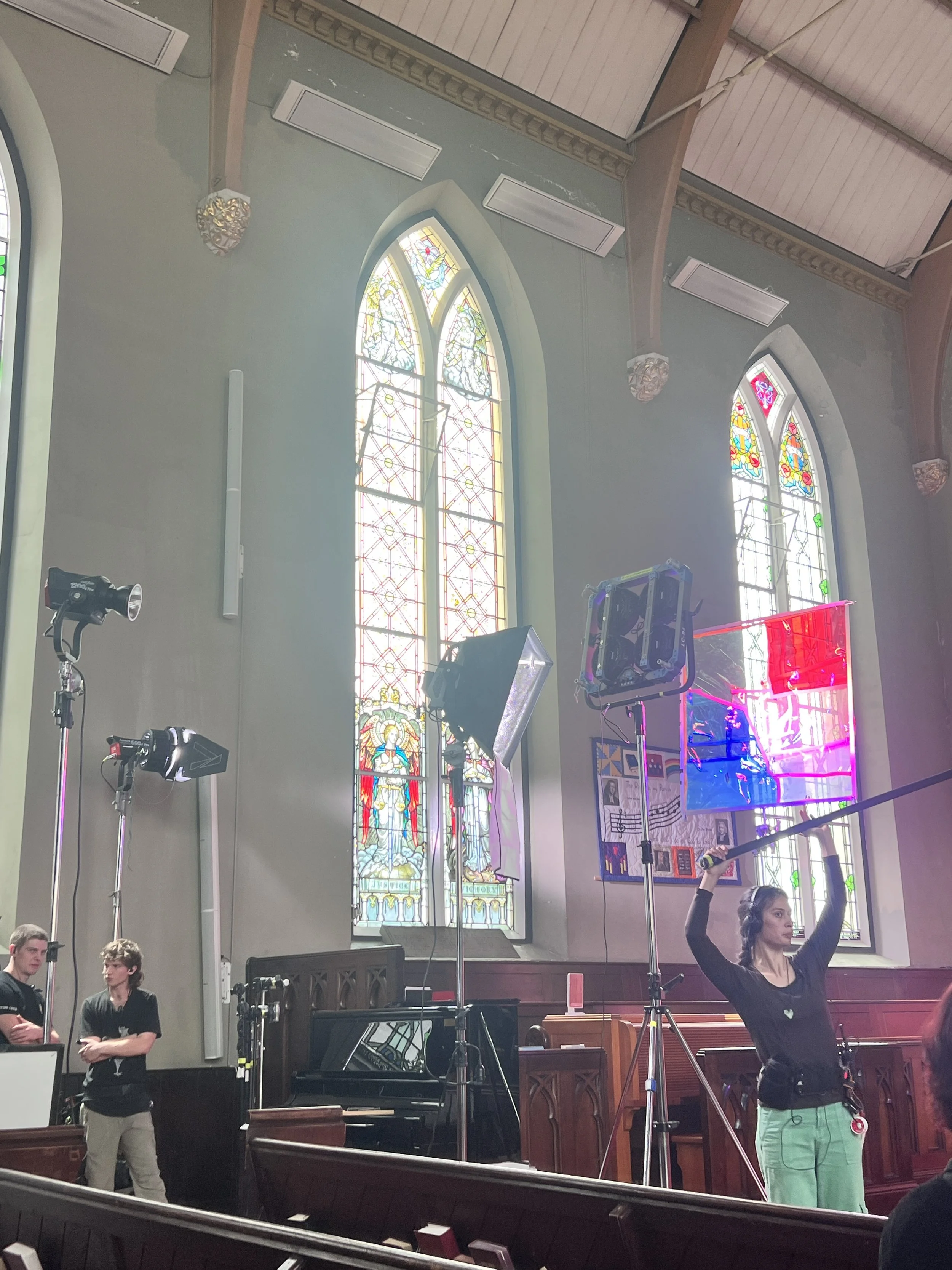 A woman in black shirt and green pants holding a long pole with a rectangular lighting panel, inside a church with stained glass windows, stage lights, and people preparing for filming or photography.