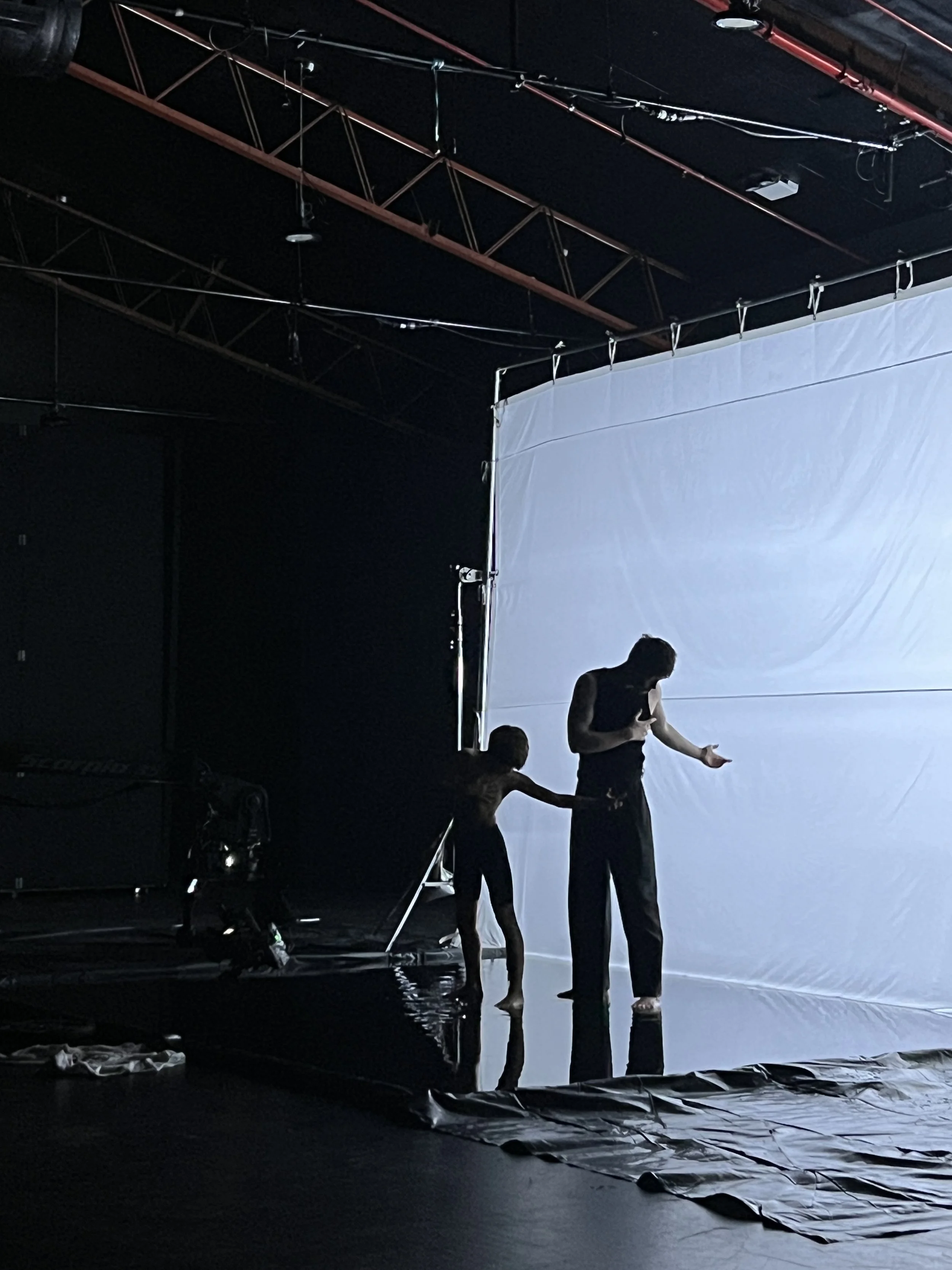 Two performers, a woman and a man, are on a stage with a white backdrop. The woman is on the left, reaching out with her right hand, while the man on the right is looking down at his hand, holding it out. The stage has black flooring with some equipm