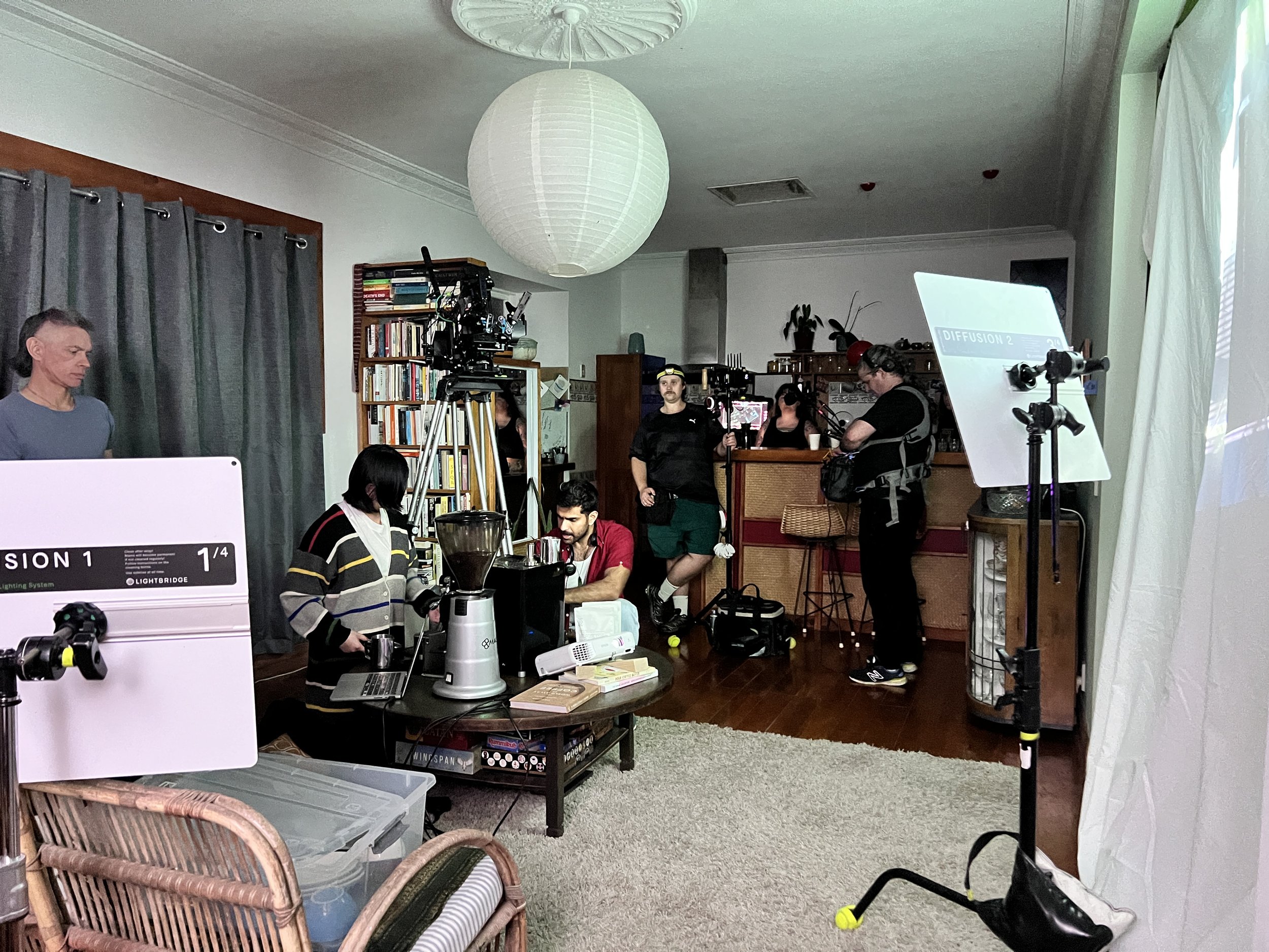 Interior of a living room set up for filming, with crew members operating lighting and camera equipment, a woman in a striped sweater preparing coffee, and a man sitting on a stool near a kitchen bar.