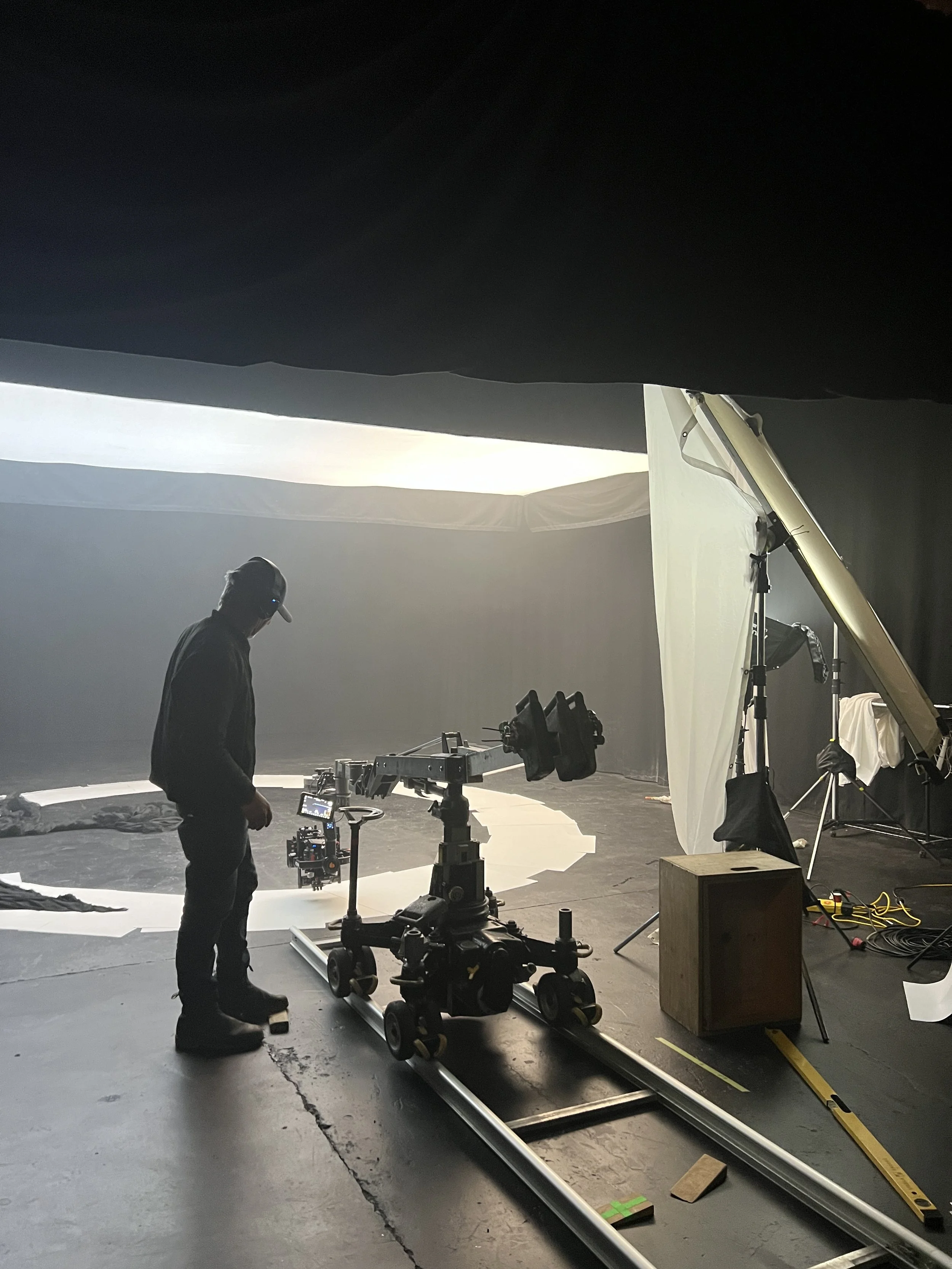 A man stands on a film set with a camera rig on a track, surrounded by studio equipment, while filming a scene in a large, dimly lit space with a white circular backdrop and white fabric overhead.
