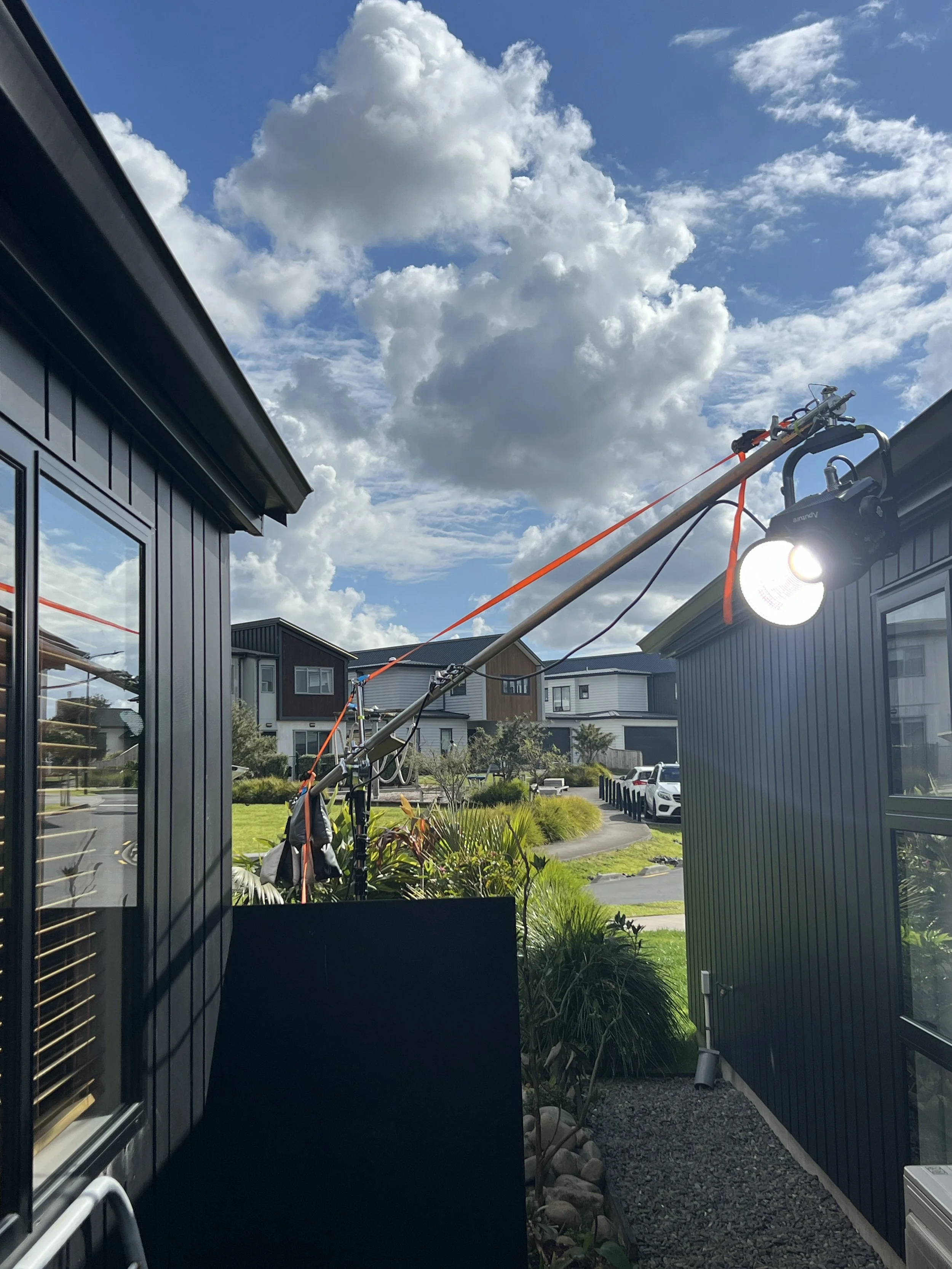 A newly installed outdoor light fixture mounted on a building with a long pole and orange straps. The pole extends between two houses under a partly cloudy sky, with a street and other houses visible in the background.