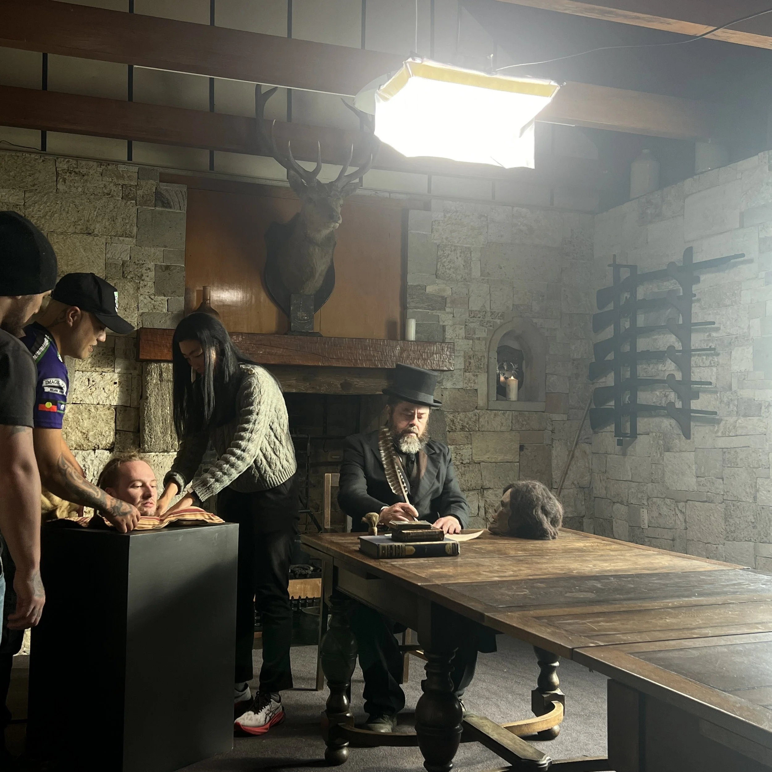 A scene recreated to look like a historical or Western setting with a man at a desk, some glasses, and books. There are several people, one appearing to examine a deceased person on a table, a mounted deer head on the wall, and a background with ston