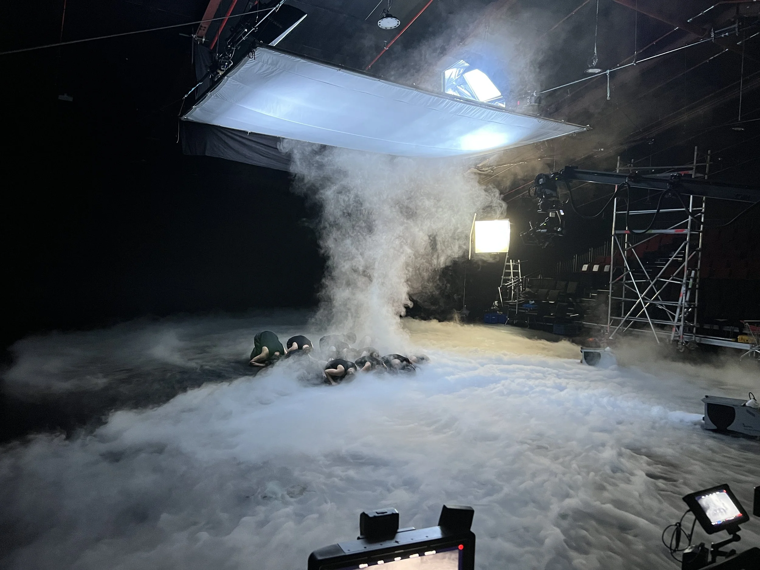 A film or stage set with a fog or smoke effect, several crew members working on the floor, large lighting equipment, and scaffolding, with a white overhead screen or reflector overhead.