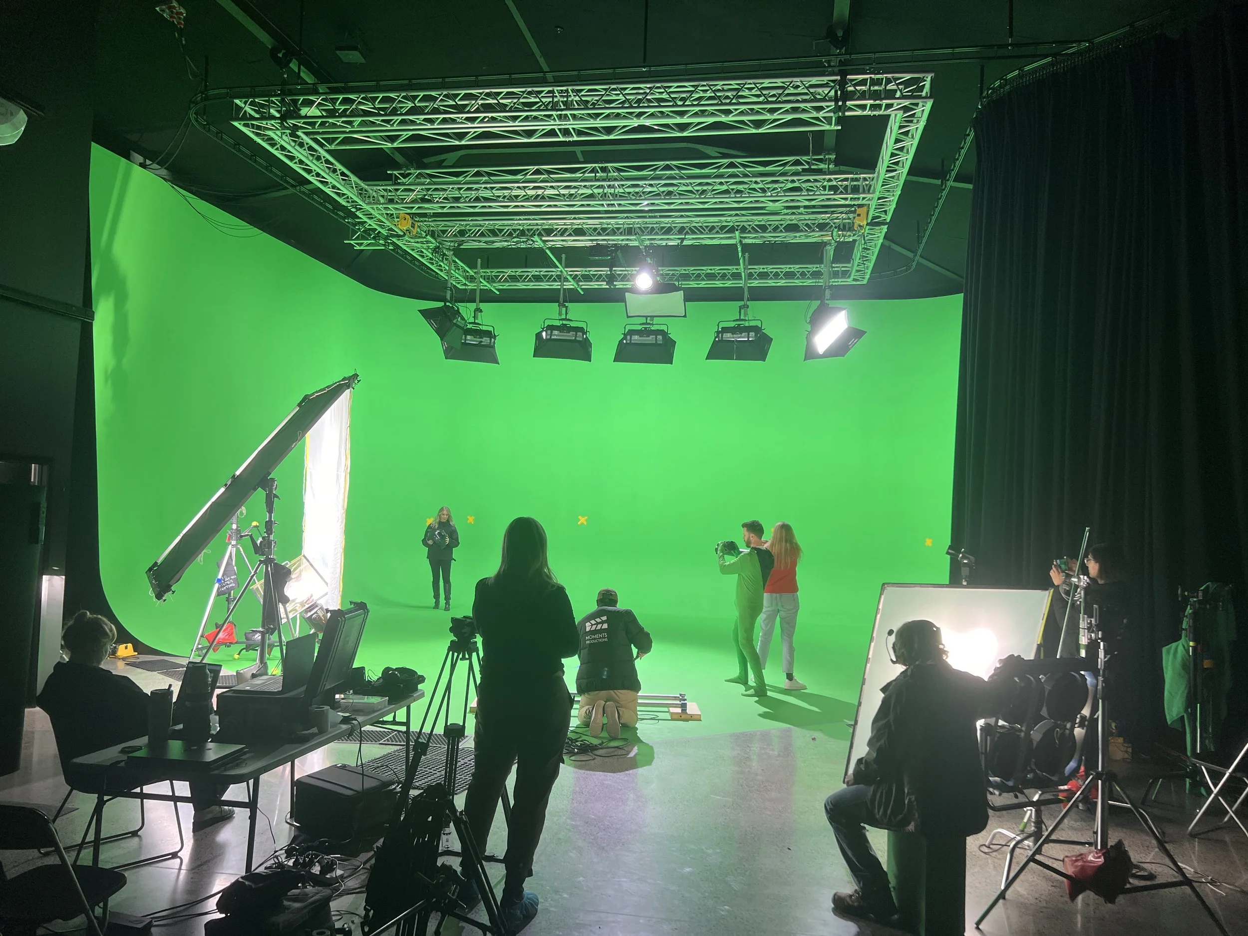 Green screen studio with crew preparing for filming, including a woman on set, photographers, and lighting equipment.