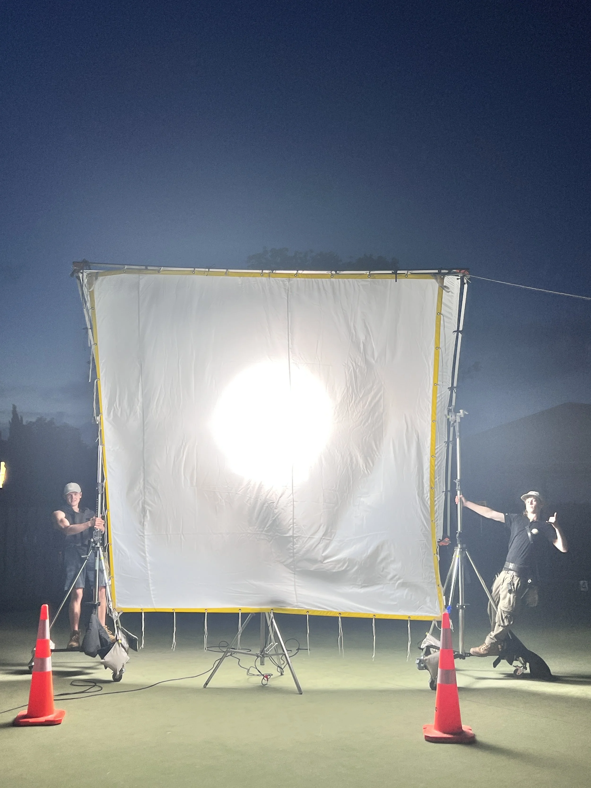 Two crew members set up a large white screen with a bright light shining through it, outdoors at night, with orange traffic cones on the ground.