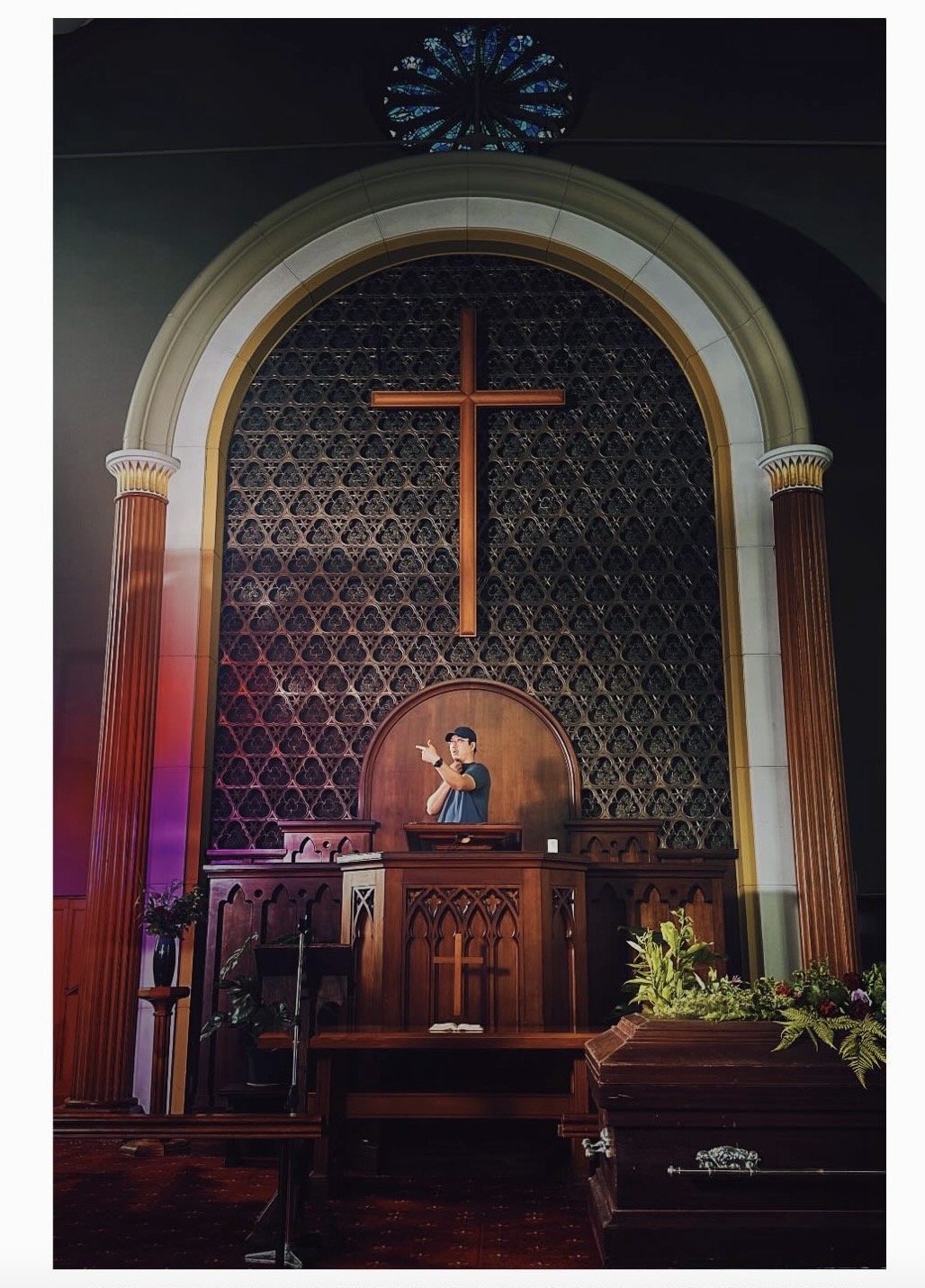 Interior of a church with a wooden altar, a large cross on the wall, and a mural of a young man pointing, surrounded by columns and decorative elements.