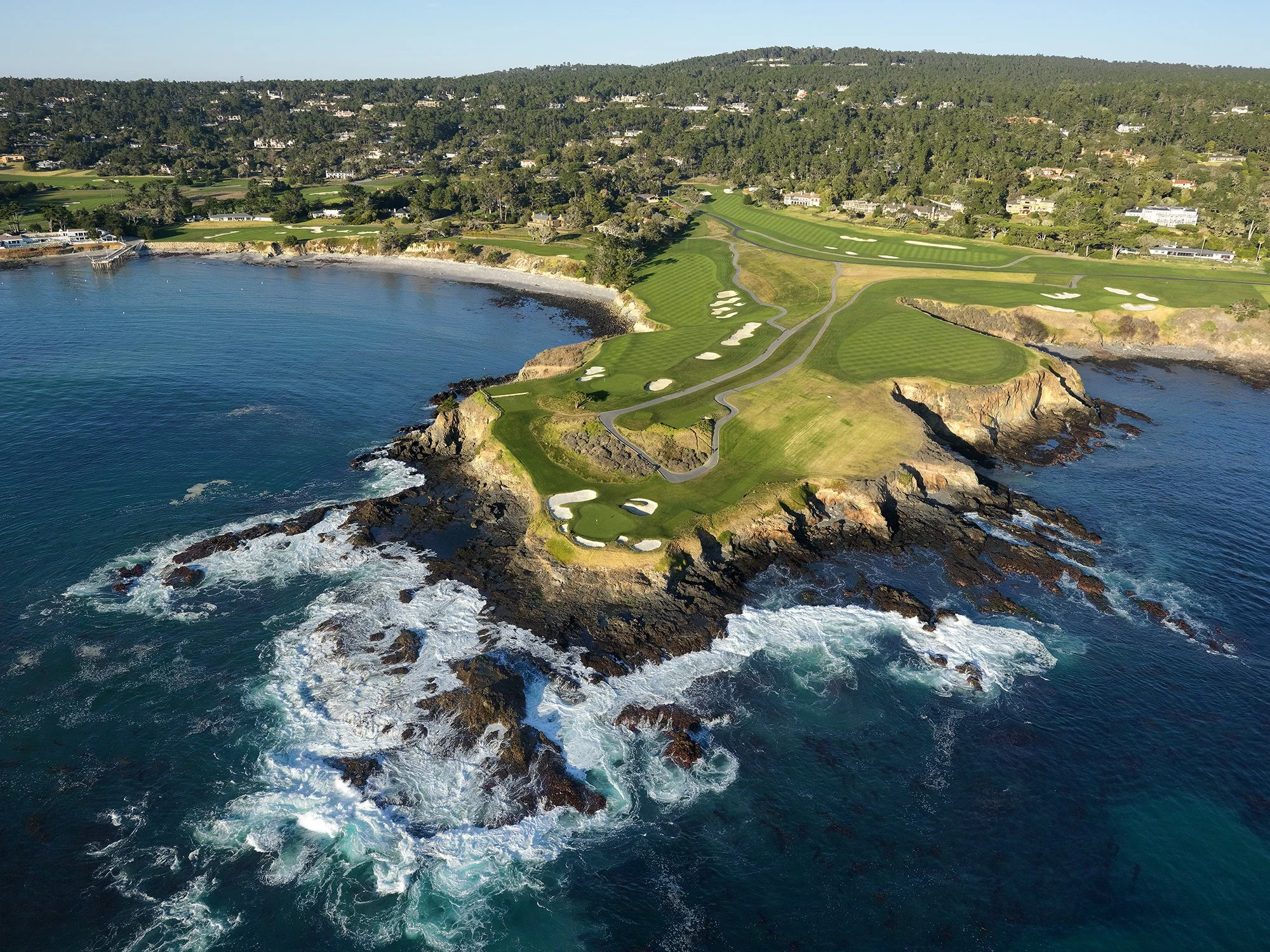 Aerial view of a coastal golf course with rolling green fairways, sand traps, cliff edges, and ocean waves crashing against rocks.