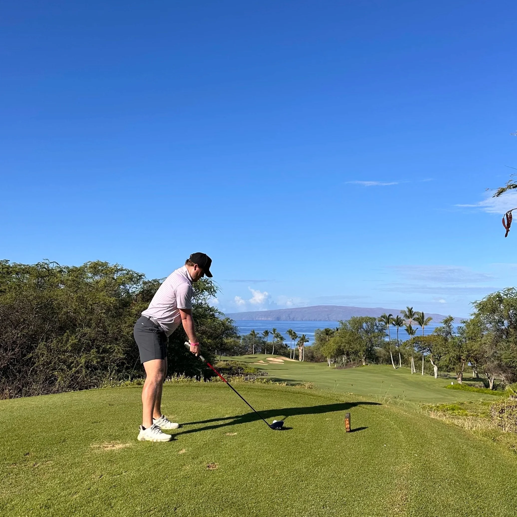 A man in a golf course, preparing to hit a golf ball using a driver club. The background shows a scenic view of water, islands, palm trees, and a mountain range under a clear blue sky.