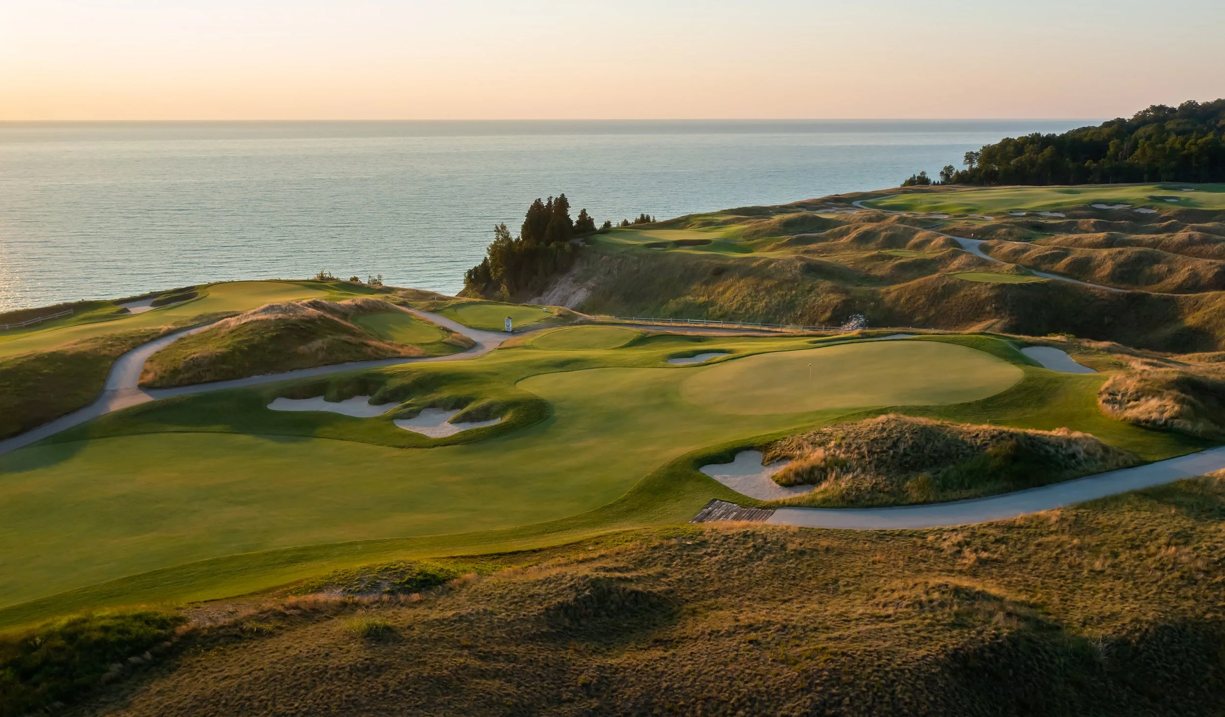 A scenic view of a golf course situated on coastal cliffs during sunset, with lush green fairways, sand bunkers, and a calm ocean in the background.