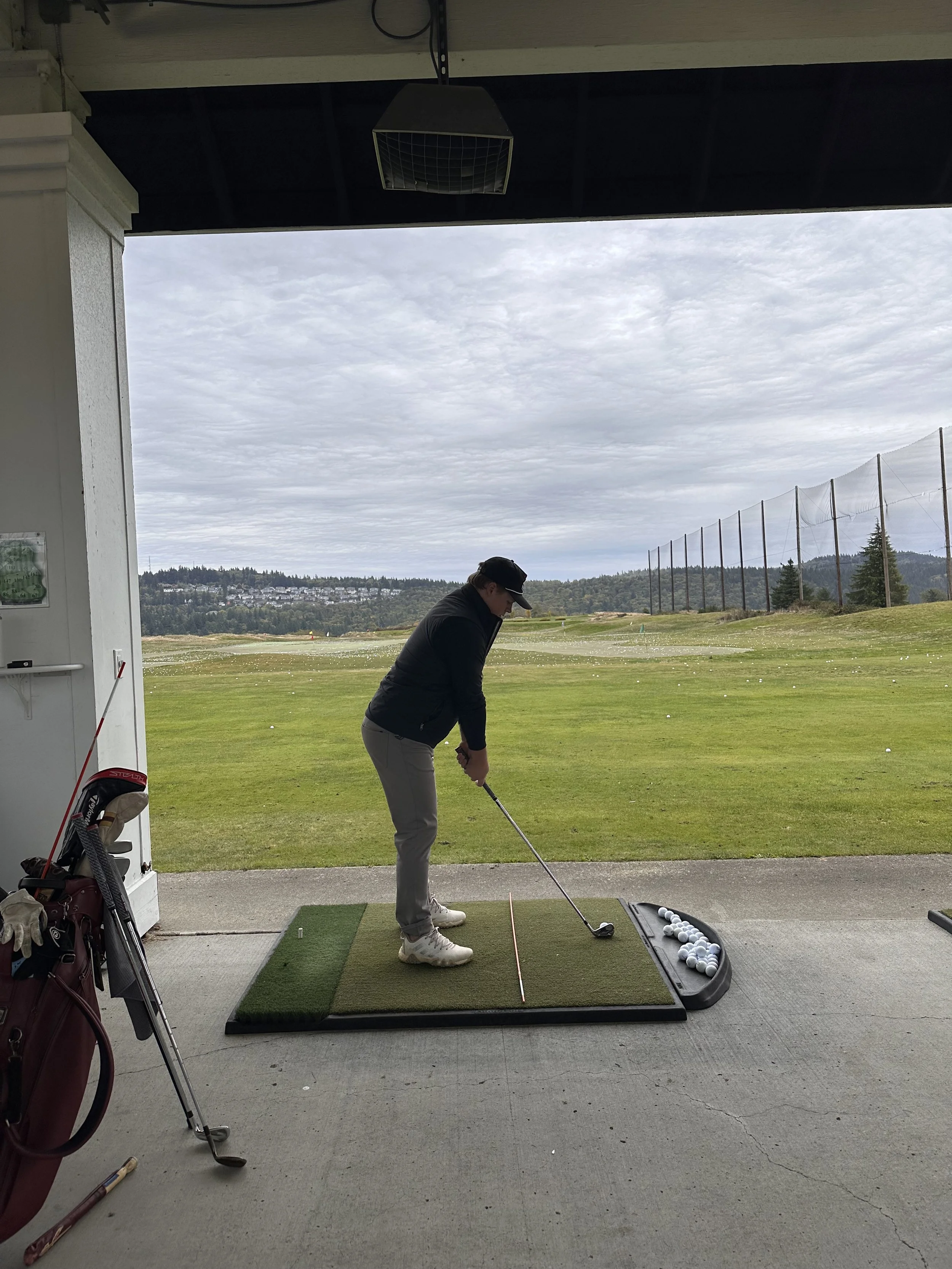 A person is practicing golf at a driving range, preparing to hit a golf ball with a club. The scene is outdoors with groves in the distance and a cloudy sky overhead.