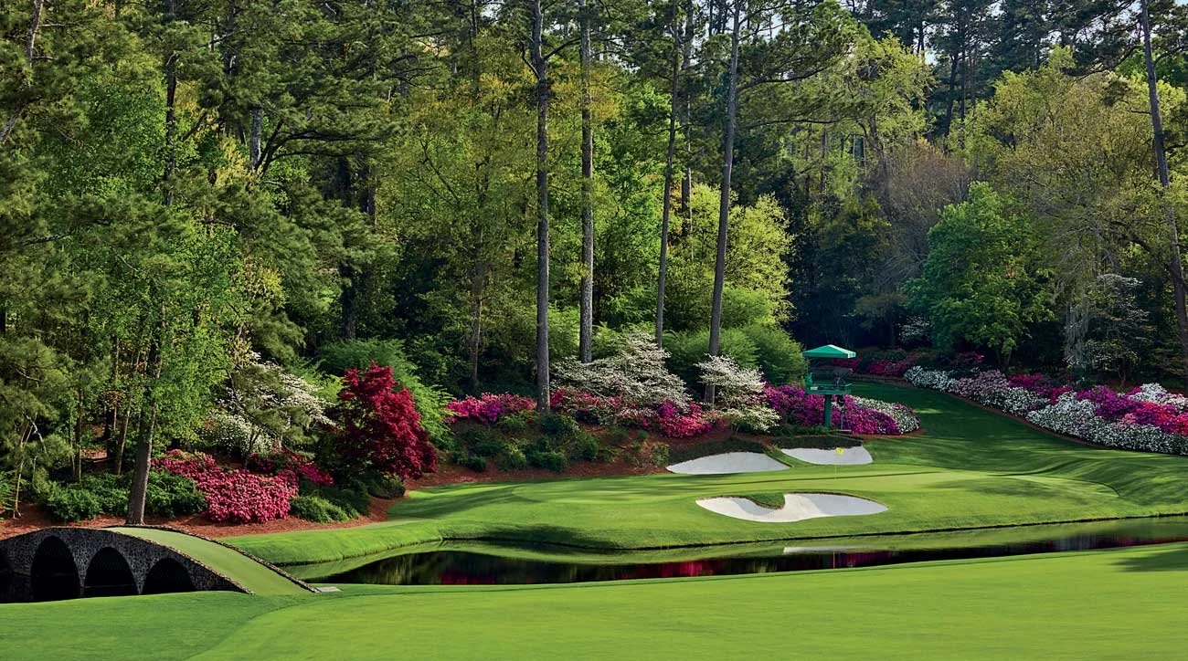 A golf course surrounded by trees and colorful blooming flowers, with a bridge over water and a small green structure in the background.
