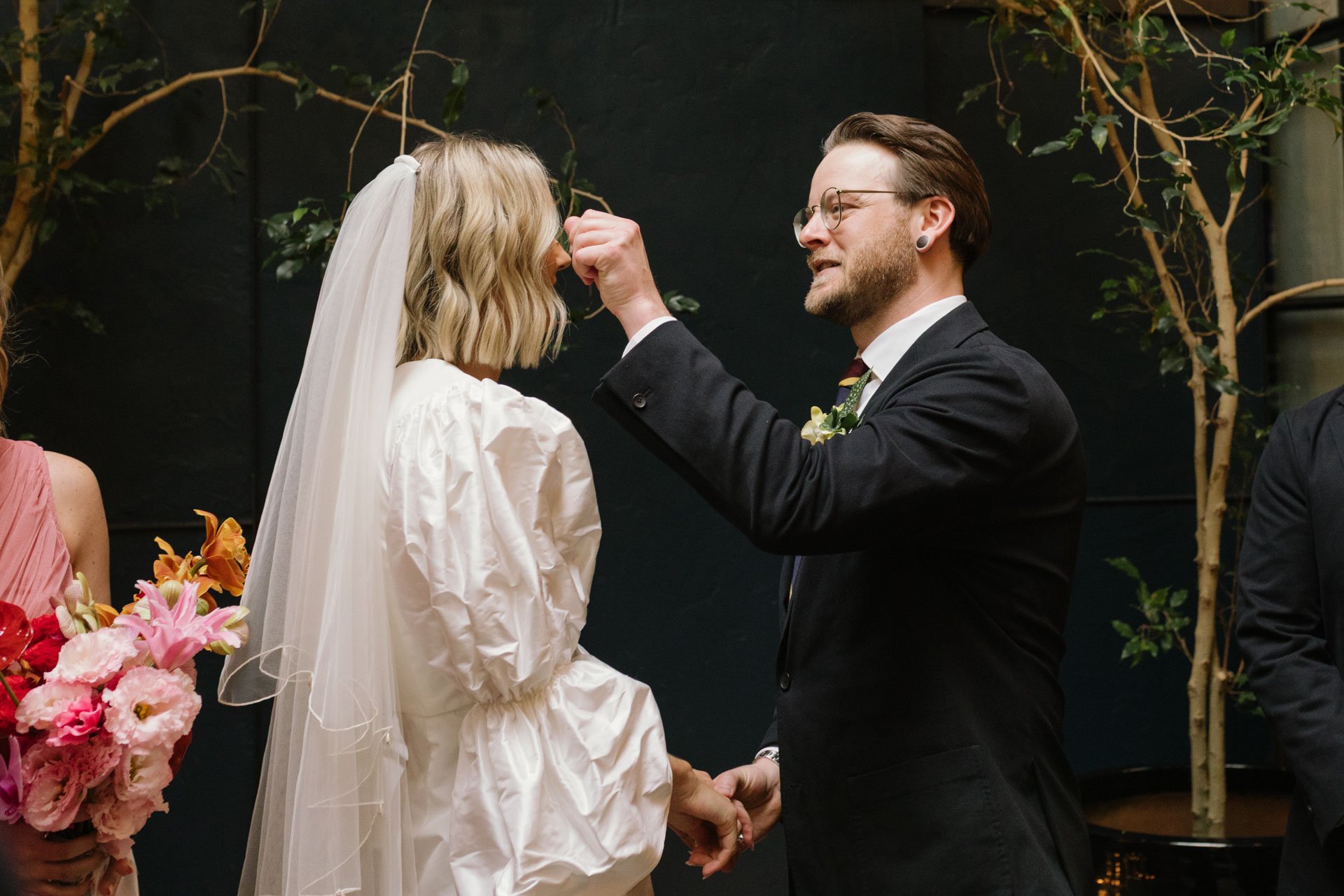 A groom is lifting the bride's veil during their wedding ceremony, holding her hands and smiling. The bride is wearing a white dress and veil, and the groom is in a black suit with glasses. There are colorful flowers in the foreground and decorative 
