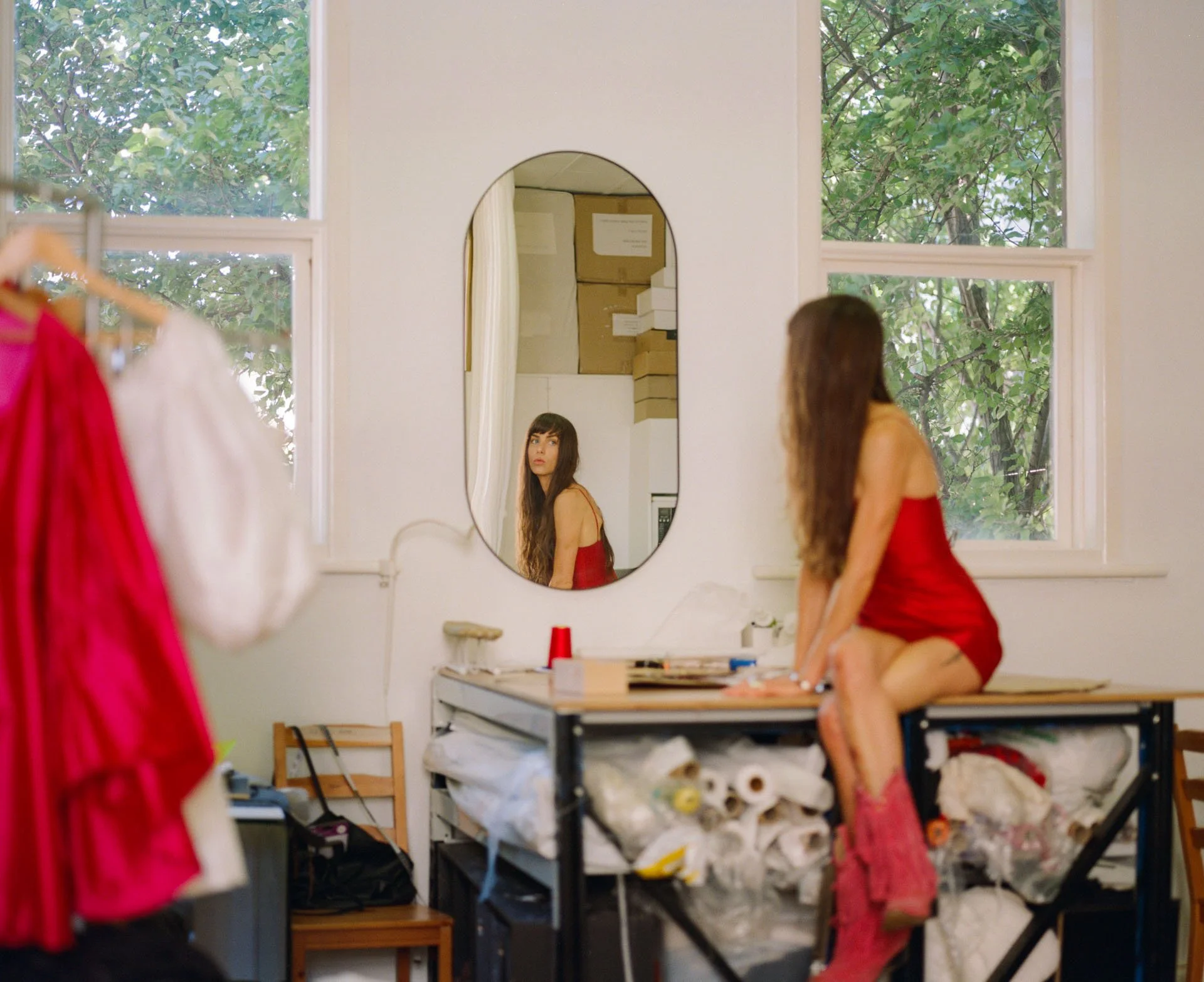 A young woman kneeling on a table in an art studio, looking at her reflection in an oval mirror on the wall, with windows showing green trees outside, and shelves with boxes in the background.