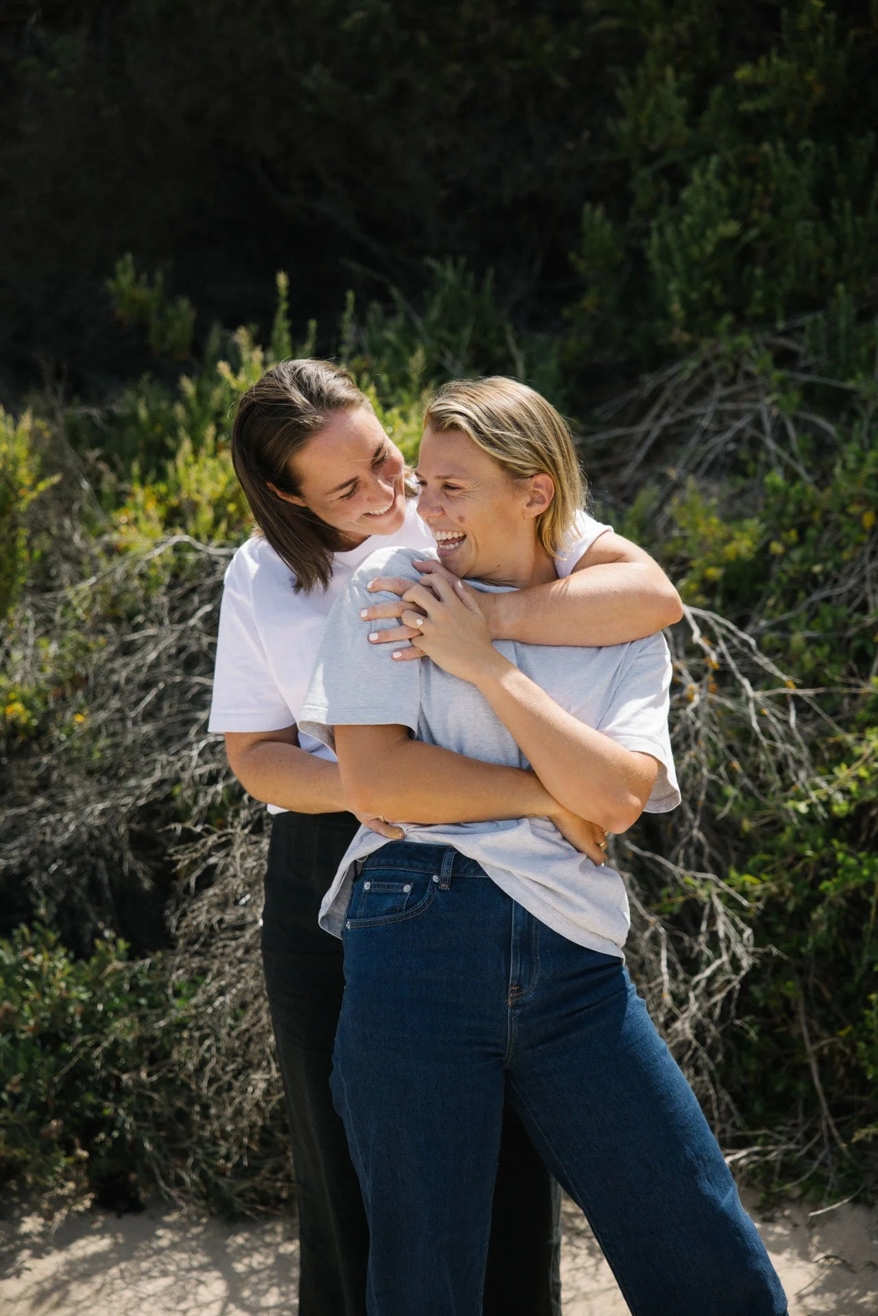 Two women embracing and laughing outdoors with greenery in the background.