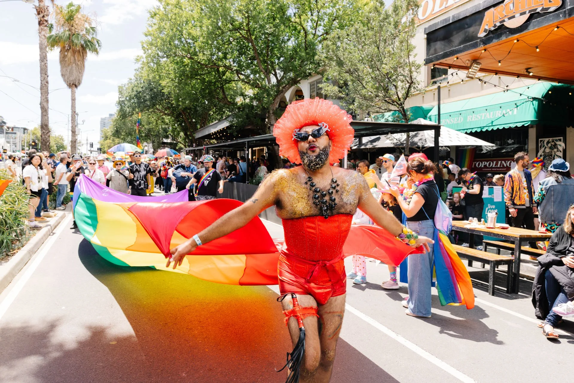 A person dressed in vibrant red outfit, with a large orange ruffled headpiece and rainbow-colored wings, parades through a lively pride parade on a sunny day, surrounded by a crowd of spectators.