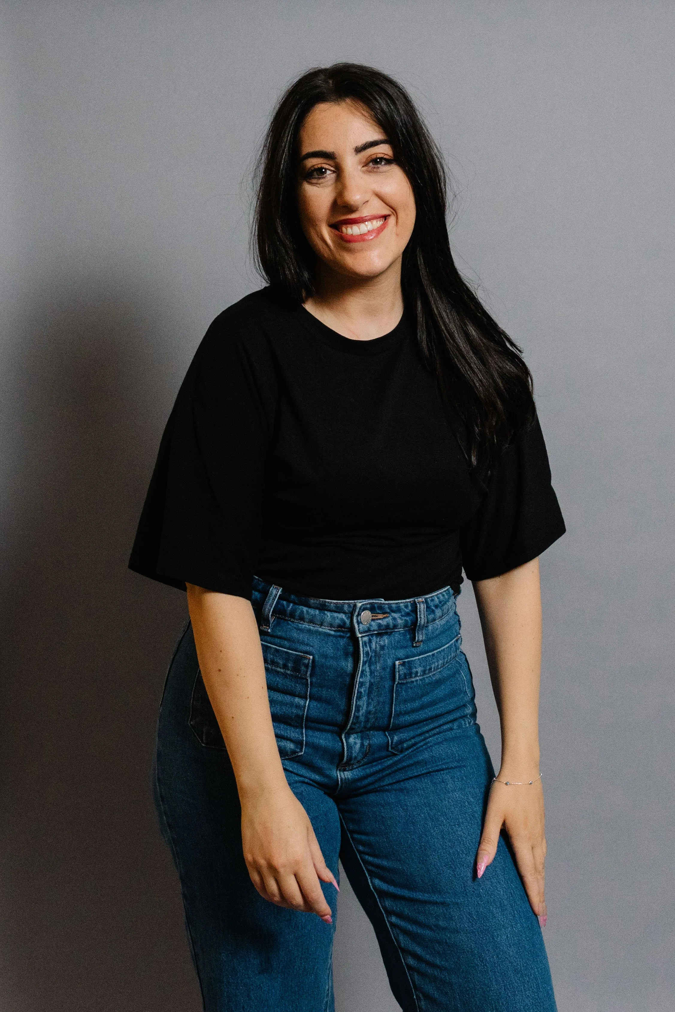 A young woman with long dark hair, wearing a black T-shirt and high-waisted blue jeans, smiling while standing against a plain gray background.