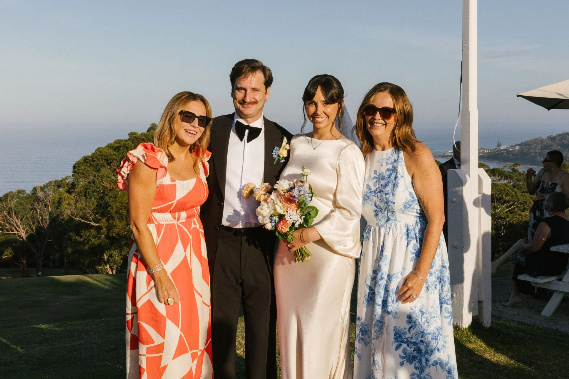Four women and one man in formal attire at an outdoor wedding ceremony, with scenic coastal background and greenery.